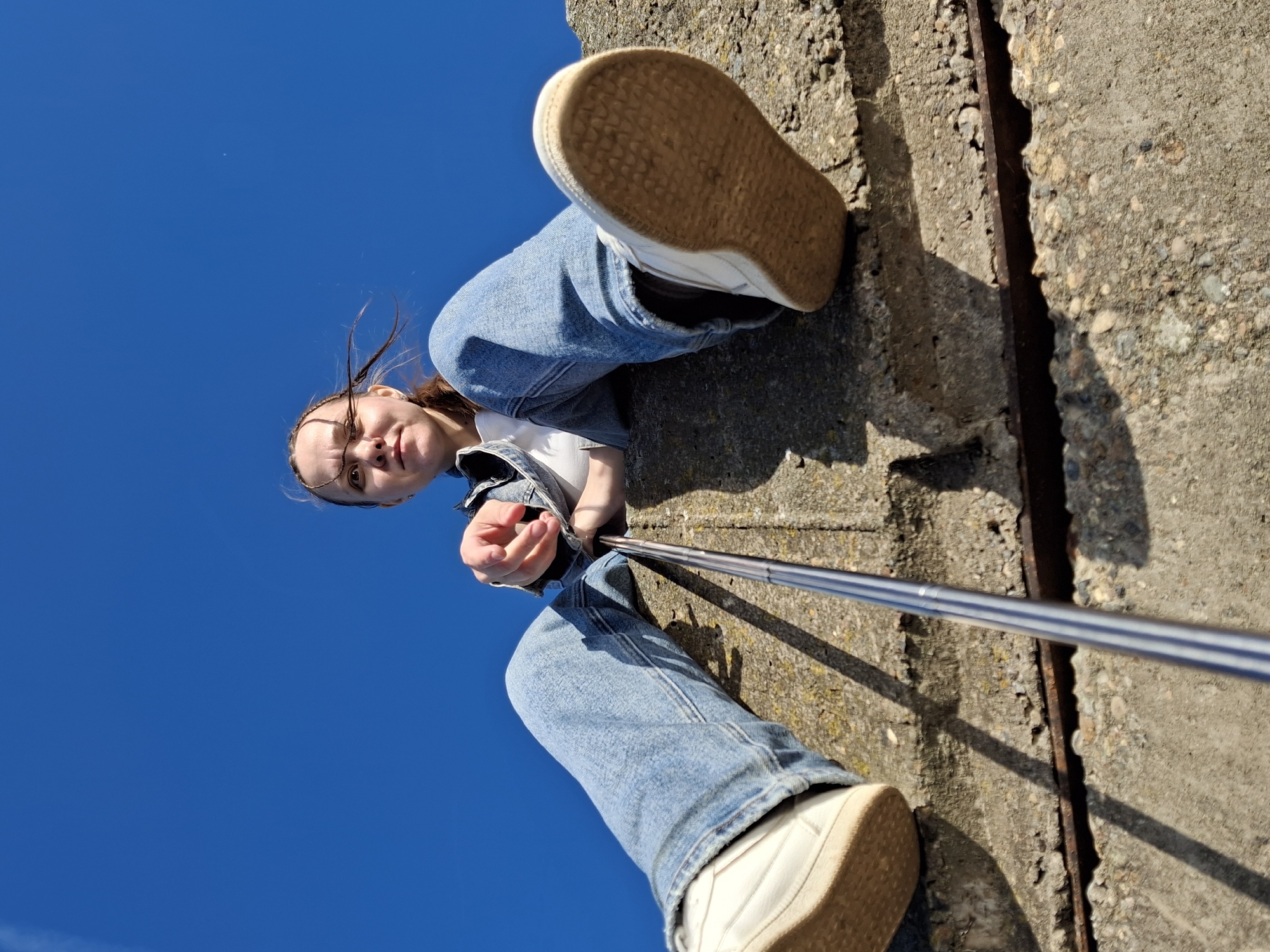 A low-angle shot captures a young person looking downwards towards the camera, holding what appears to be a selfie stick. The subject, a young female, is dressed in a white t-shirt, blue denim jeans, and white sneakers with distinctive beige soles. Her brown hair is slightly wind-blown, and she maintains a neutral, direct gaze.
The individual is positioned against a rugged concrete surface that dominates the right side and foreground of the image. This concrete structure, possibly a wall or ledge, shows signs of age with its coarse texture, visible aggregate, and cracks. A rusty metal bar is embedded lengthwise within a crevice in the concrete. The lighting is bright and clear, with strong, dark shadows cast across the concrete, indicating direct sunlight on a sunny day.
The expansive background is a vivid, cloudless deep blue sky, filling the left and upper portions of the frame. The perspective suggests the camera is placed very low, looking up at the person who might be standing or sitting on an elevated structure. The setting conveys an outdoor, possibly urban or industrial environment, consistent with areas found in Chaikovsky, Russia. No visible text is present in the image.
