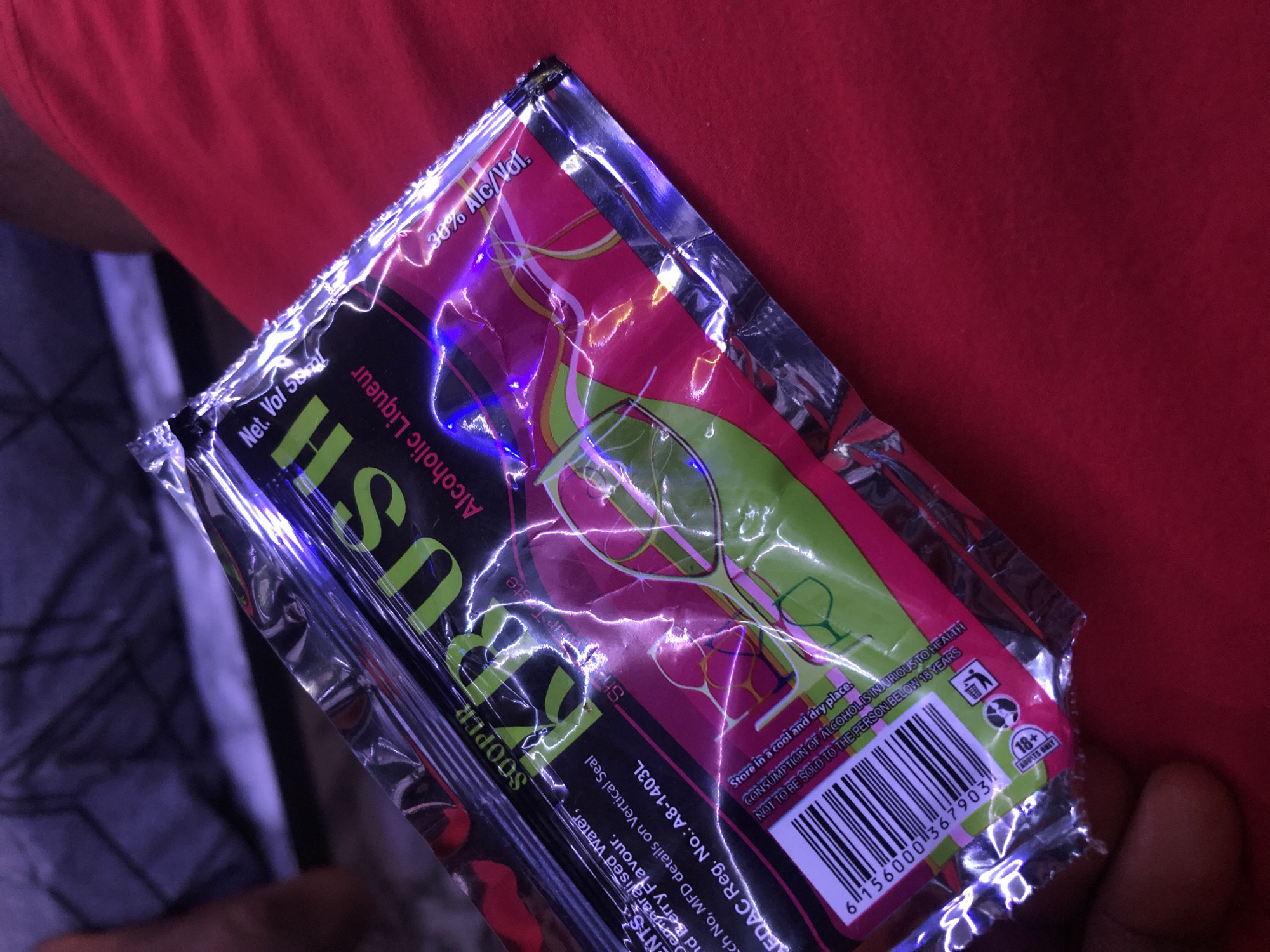 A close-up shot shows a foil packet for an alcoholic liqueur product, labeled "KRUSH," with a net volume of 55ml and 38% alc/vol. The packet is predominantly pink and green with silver accents and displays a barcode and "ADULTS ONLY 18+" icon. The packet is resting on a red fabric surface, possibly clothing. The background is out of focus, showing a person in dark attire and a wall with a textured surface and purple lighting. The image appears to be taken in a domestic setting, as there are no specific indicators of New Stadium, Nigeria.