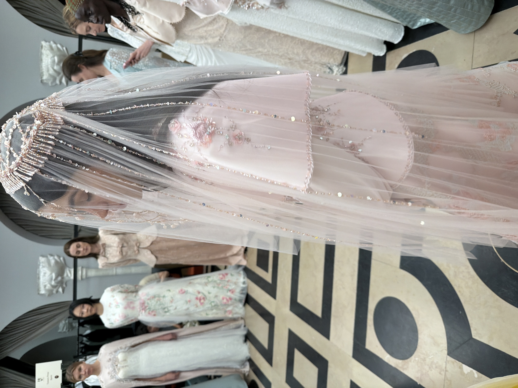 The image captures a scene indoors, potentially within a fashion show or presentation venue. The floor is covered in a bold, geometric pattern of alternating black and off-white shapes, predominantly consisting of interlocking "C" shapes and circles.
Foreground: A woman wearing a pink dress with a sheer, heavily embellished veil. The veil has a beaded and sequined edge, and more scattered sequins throughout. The dress has floral embroidery.
Midground: Behind and to the left of the veiled woman are several other individuals in what appears to be bridal wear or formal dresses. The dresses vary in style and color, with some featuring floral patterns, lace, and other embellishments. These women are standing still.
Background: To the left, there is what seems to be an architectural detail, possibly a section of wall or a decorative column topped with a plaster bust. A sign is visible on the bottom left corner.
Environment: The lighting appears to be controlled, likely from stage or gallery lighting.
The location is in Paris, France.