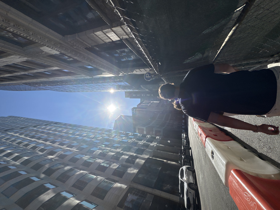 This image depicts a pedestrian-level view looking south down Congress Street in the Financial District of Boston, Massachusetts, United States. The clear blue sky shows a prominent sunburst with lens flare, indicating a bright, sunny day.
On the left side of the street, the modern, multi-story skyscraper at 100 Federal Street rises, characterized by its distinctive curved, reflective glass facade and numerous windows. Adjacent to the street, a line of interconnected red and white plastic construction barriers separates the roadway from parked vehicles. Several cars are visible, including a white SUV parked closest to the foreground.
On the right side, an older, tall building with a dark, ornate stone or brick facade features classical architectural details, including cornices and multiple rectangular windows. A dark mesh-covered chain-link fence runs along the sidewalk in front of this building, likely indicating a construction or renovation site. A blue circular "P" parking sign is affixed to a pole or the fence.
In the foreground, a male individual is walking away from the camera. He has short, curly brown hair, wears a dark short-sleeved polo shirt, and light-colored shorts or pants. His left arm is visible, showing a bracelet or watch. A second person is partially obscured behind his left shoulder. The street is asphalt, and visible shadows are sharp due to the direct sunlight.