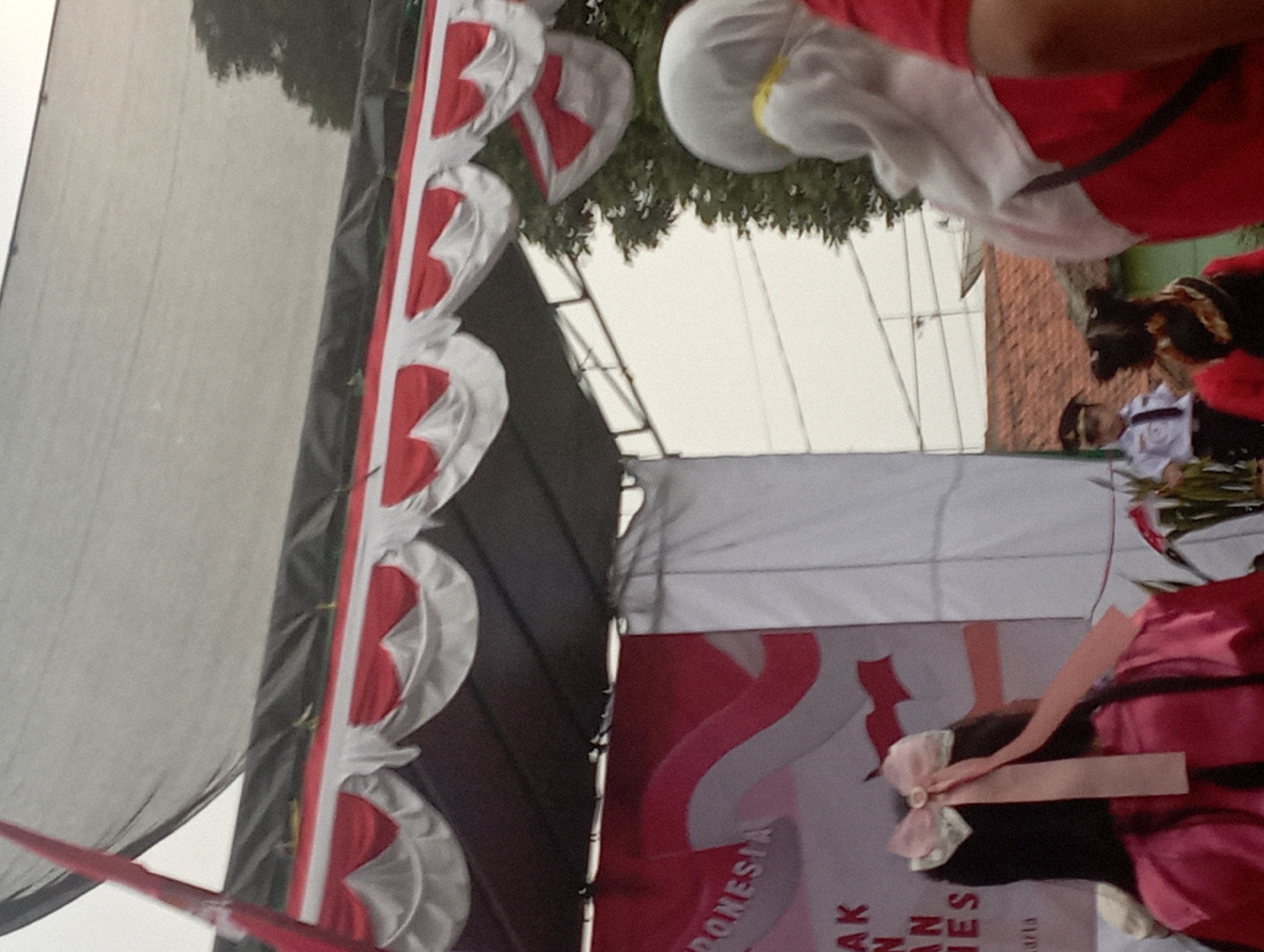 In a mid-range outdoor shot, the image captures an event with a stage decorated in red and white, resembling the Indonesian flag. A black canopy stretches over the stage, with festive red and white fabric drapes hanging below. To the left, a large banner displays red and white wavy patterns, with text that appears to say "INDONESIA". In the foreground, a person with long dark hair is wearing a bright pink satin outfit and a pink bow with two long ribbons on their head. Further into the scene, another person wearing a white hijab and red garment is partially visible on the right. In the middle ground, a child in a white uniform with a black tie and cap is standing, possibly participating in the event. Behind them, more individuals are present, and glimpses of trees and rooftops suggest an urban or semi-urban setting in Purwakarta, Indonesia. The sky is overcast, indicating a potentially cloudy day.