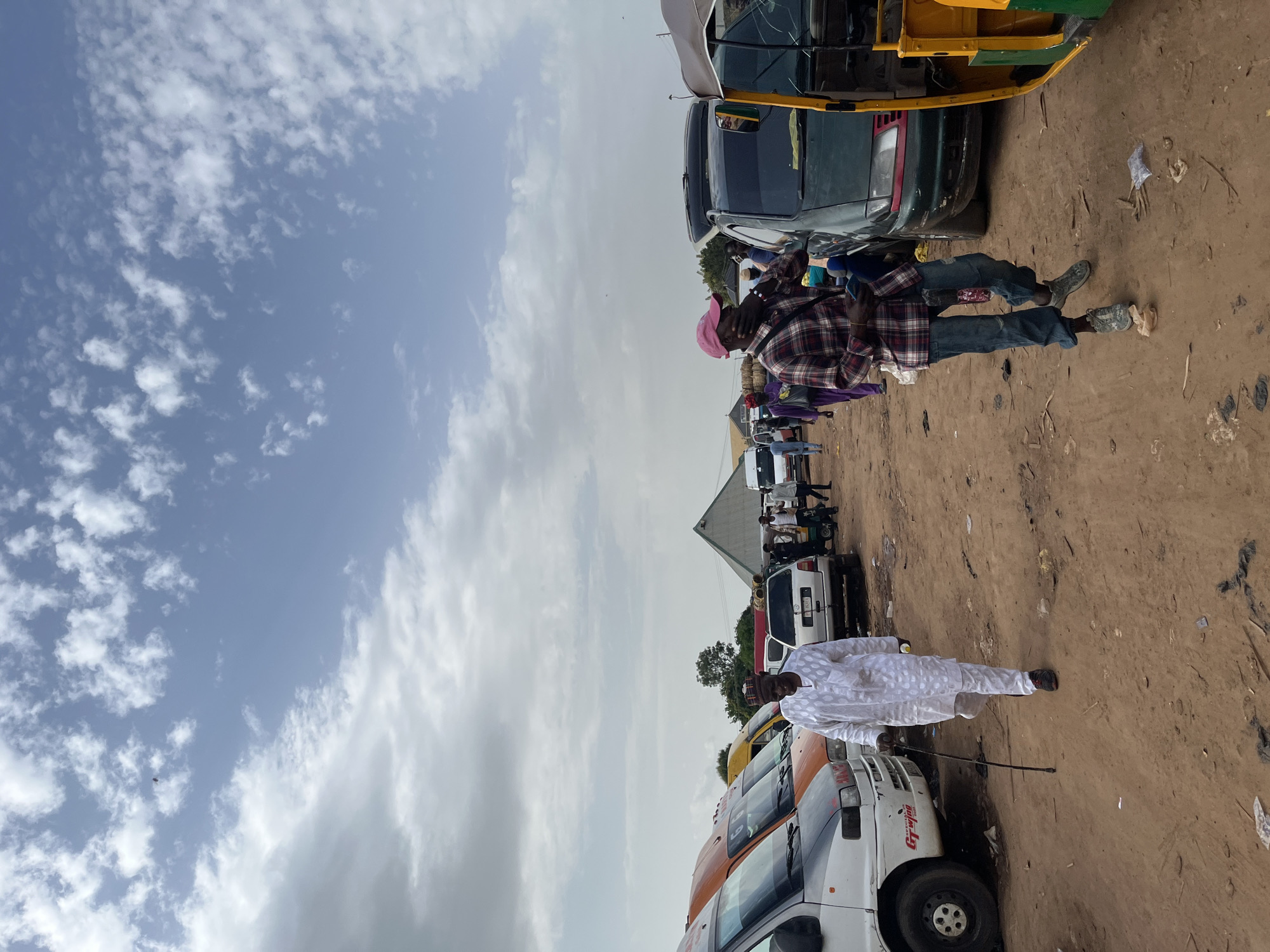 The image depicts a dusty, outdoor scene with several vehicles and people in what appears to be a transportation hub or market area in Rafin Par, Nigeria. The sky is partially cloudy with blue sky visible.
In the foreground, a man wearing a white traditional outfit and a cap walks from left to right, leaning on a dark walking stick. He is looking towards the viewer. To his right, further into the scene, another man is walking away from the camera, wearing a plaid shirt, blue jeans, a pink cap, and a dark mask. He is holding a phone in his left hand.
Several vehicles are parked in the scene. On the left, a white van with orange accents is visible. Behind it, several other vans and a green car are parked. In the far right of the image, part of a yellow and green auto-rickshaw is visible.
Other people are scattered throughout the scene, some walking or standing near the vehicles. The ground is covered in dirt and debris, indicating a somewhat unpaved area. In the background, a large tent-like structure with a pointed roof is visible. The overall impression is of a busy, informal gathering point for transportation and commerce.