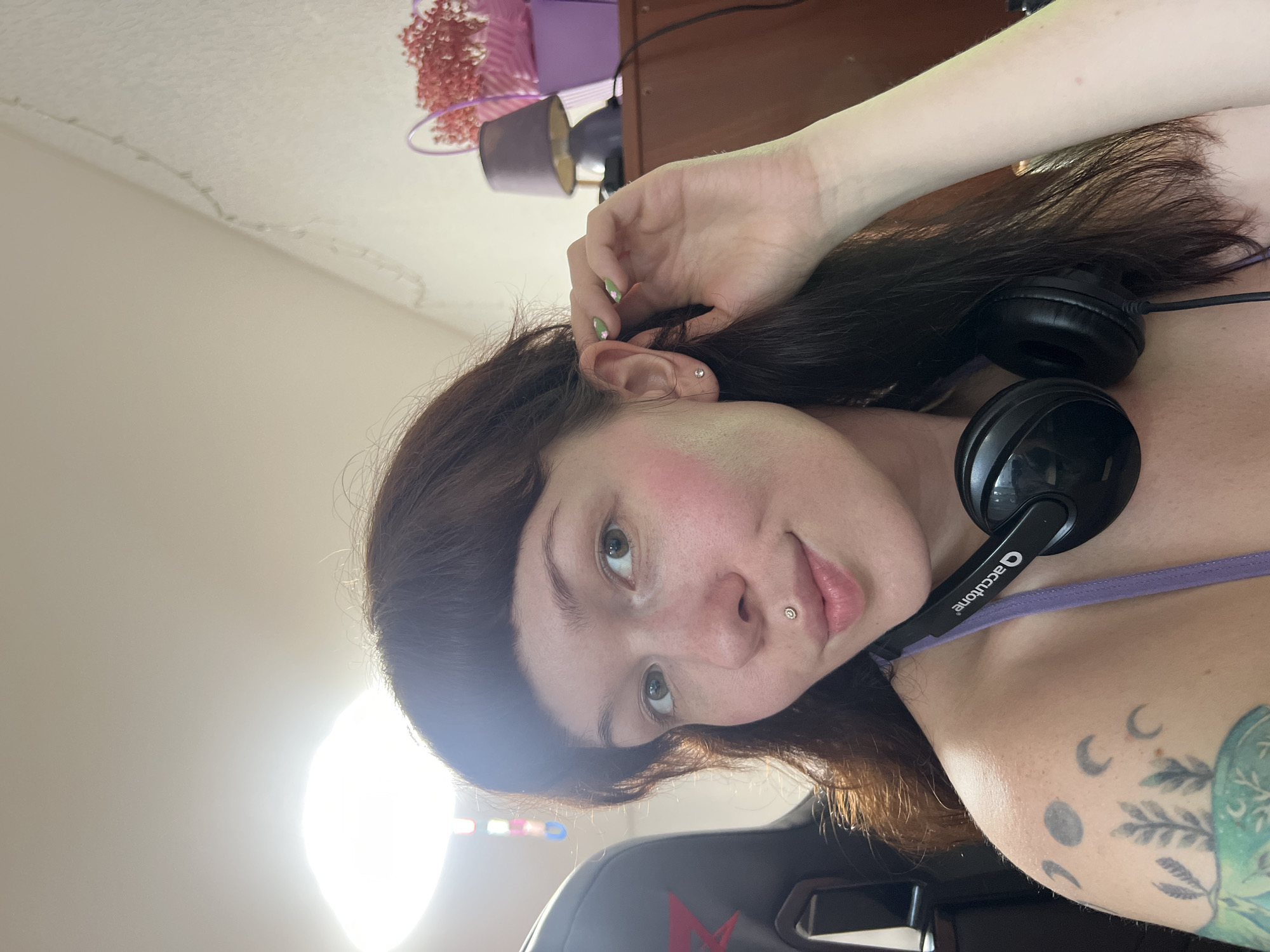 A close-up, indoor shot focuses on a young woman with dark brown hair and bangs, a lip piercing, and a nose stud. She is wearing a purple top and has headphones around her neck. Her right hand is touching her ear, and her left arm, visible in the lower left corner, is tattooed with celestial and plant imagery. The background includes a gaming chair, a shelf with a purple lamp and a basket of dried flowers, and a wall adorned with a string of lights. The lighting suggests an artificial light source is present, possibly a ring light. There is no indication of the city of Chaikovsky, Russia, in the image.