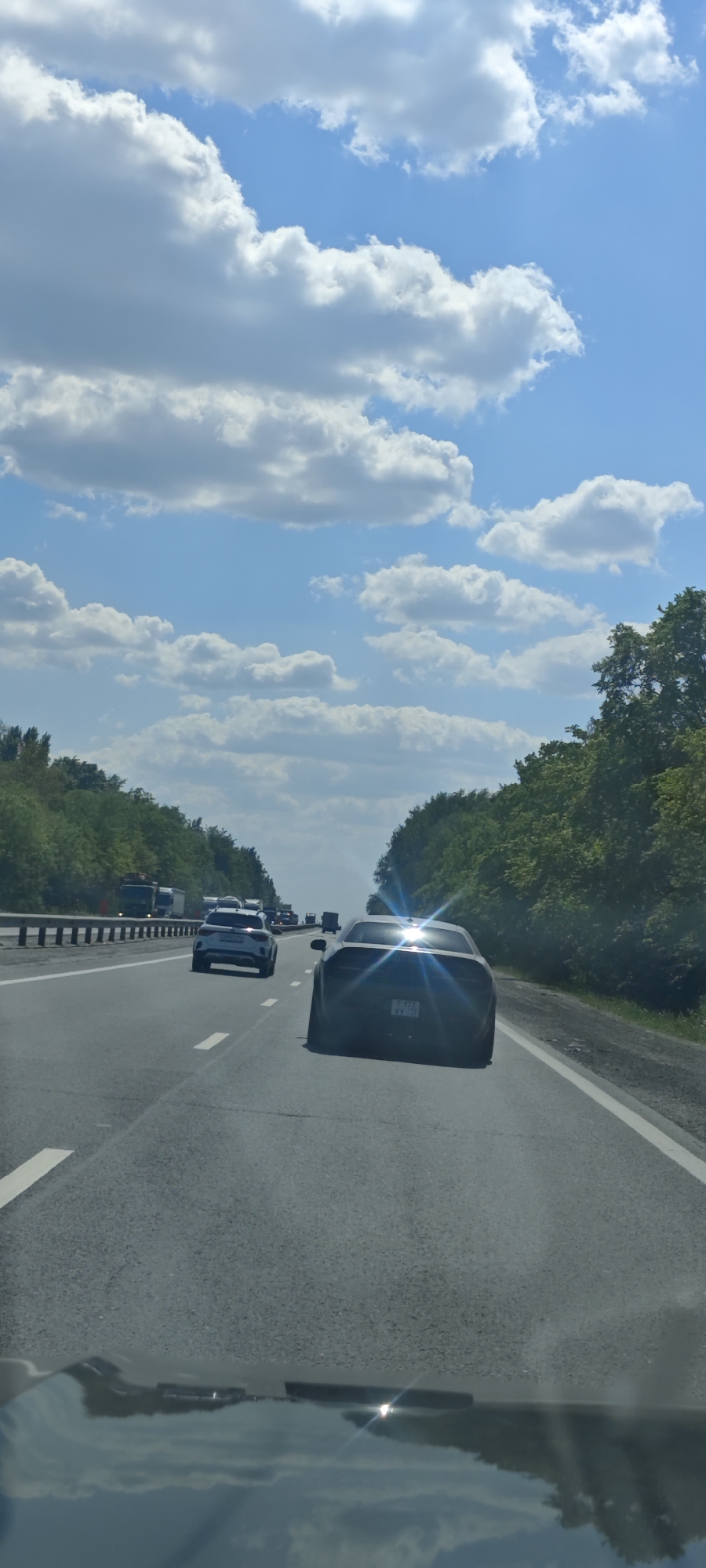 The image shows a view from inside a car on a highway, looking forward. Other cars are visible ahead, with the closest car being a dark-colored sports car. The highway is bordered by trees on both sides. Above, the sky is blue with numerous white clouds. The city of Bely Klyuch, Russia, is not identifiable from the image.