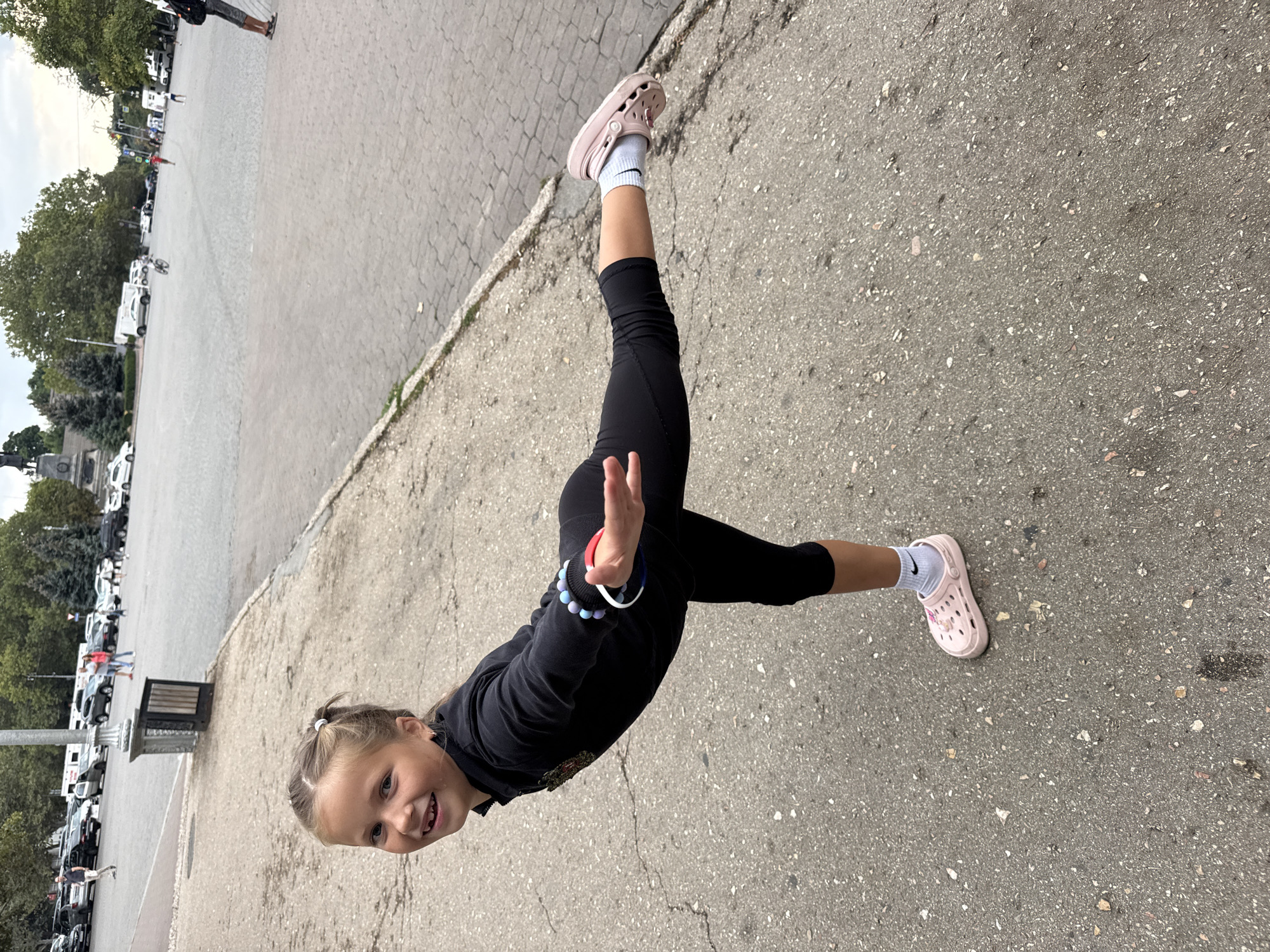 A young girl with blonde hair in a ponytail is standing on one leg in a dancer's pose on a cracked asphalt sidewalk. She is wearing a black long-sleeved shirt, black leggings, white socks with a Nike logo, and pink Crocs. She is smiling at the camera and has her arms outstretched, with one hand reaching towards the viewer. The background shows a paved plaza with cars and people in the distance, indicating an urban or public area. The sky is overcast.