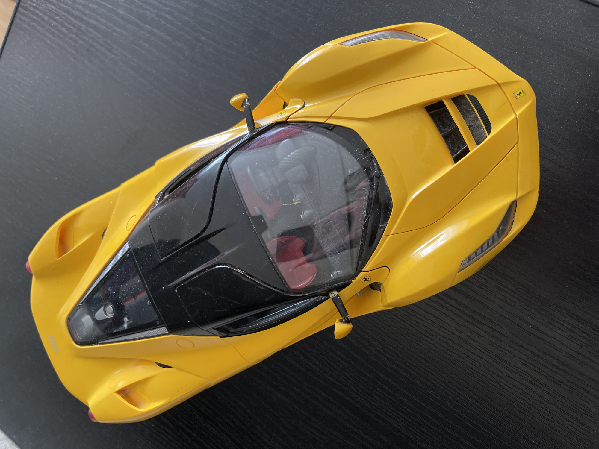 A vibrant yellow scale model of a Ferrari hypercar, resembling a LaFerrari, is captured from a top-down, slightly angled perspective. The model rests on a dark, textured surface, likely a wooden table or desk. The car features a sleek, aerodynamic design with a black-tinted canopy revealing a detailed interior with prominent red seats, a dashboard, and steering wheel structure. Small Ferrari "prancing horse" logos are visible on the rear engine cover on the right side and near the base of both black rearview mirrors. The word "FERRARI" is faintly embossed on the front hood.
Notable details include realistic air vents on the rear engine cover, clear plastic headlights and taillights, and intricate design lines throughout the body. The model shows some signs of use, with visible dust and minor scratches on its surfaces, particularly on the windshield. The overall scene is a static display of the model car under bright, even lighting, suggesting an indoor setting. No activity is taking place. The object is located in Moscow, Russia.