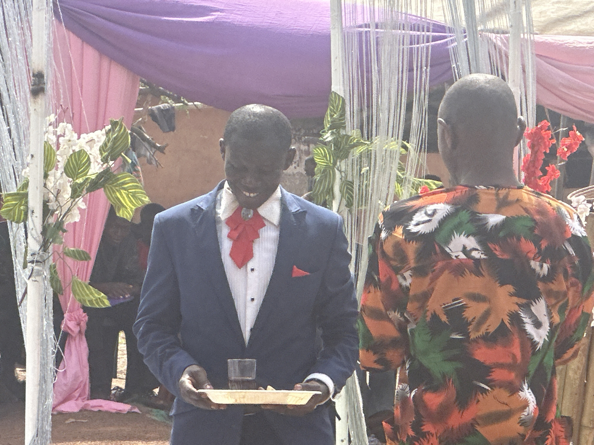 The image presents two men in an outdoor setting in Unknown, Unknown. One man, facing the camera, is dressed in a dark blue suit, a white shirt with a red bowtie, and a red pocket square. He is holding a rectangular wooden tray with a glass of dark liquid and some food on it. The other man, positioned with his back to the camera, is wearing a colorful shirt featuring a pattern of orange, brown, green, and white. The background features floral arrangements and drapery in shades of pink and purple.