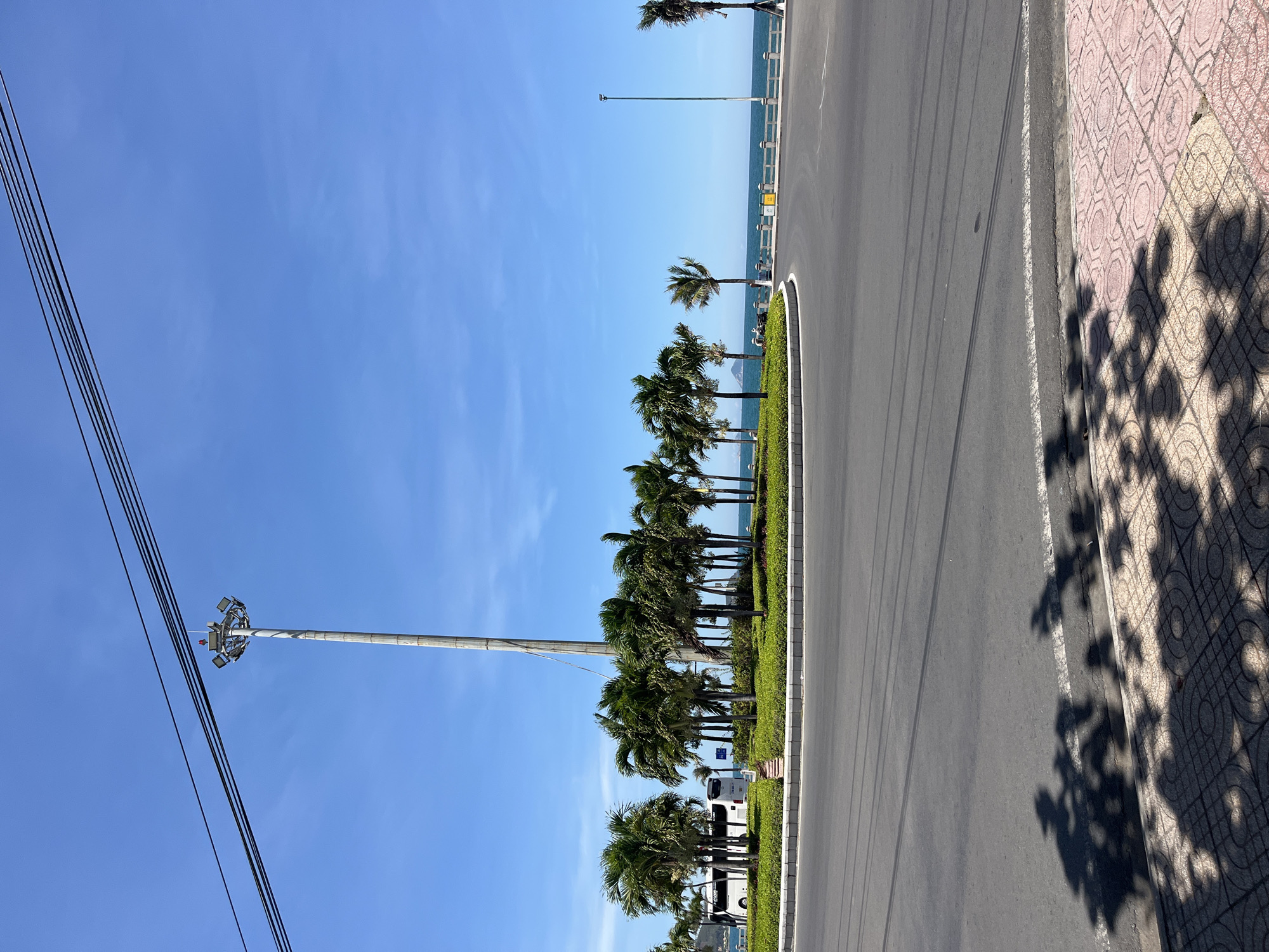 The image depicts a sunny scene in the city of Bac Nha Trang Ward, Vietnam. A street runs along a coastline with palm trees separating it from the beach. A tall light post with attached power lines is visible on the left. A sidewalk with patterned tiles runs along the right side of the road, marked with shadows from trees. In the distance, the ocean meets a clear blue sky. Several tour buses parked alongside the street, near palm trees.