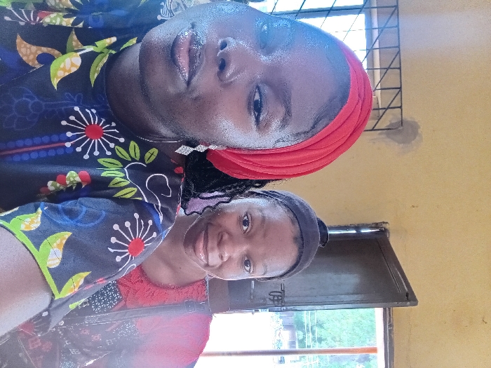 The image depicts two adult females positioned indoors in the city of Ejule, Nigeria. The foreground individual, dark-skinned, wears a red head covering, a multi-colored patterned shirt, and a silver-toned dangling earring on her visible left ear. Her lips appear glossy, and her gaze is directed towards the camera. Behind her and to the right, a second dark-skinned female is partially visible, wearing a dark headband and a reddish top, presenting a slight smile. The indoor background consists of light yellow-painted walls, one showing a grey plaster repair patch. To the left, a window with a dark metal security grille is visible, allowing light into the room. To the right, a dark brown door stands ajar, revealing an outdoor scene with green foliage and ambient light beyond.