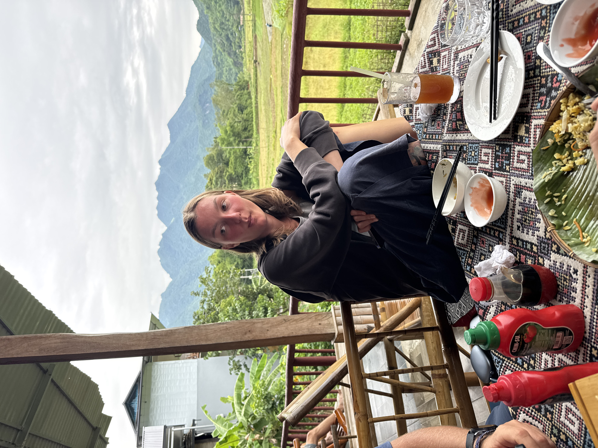 A young woman is seated at a table outdoors with a scenic mountainous landscape in the background. The table is covered with a patterned cloth and set with plates of food, bowls, chopsticks, and drinks. The woman is wearing a dark hooded sweatshirt and dark pants, and she is looking towards her right. The setting appears to be a restaurant or cafe with a view of rice paddies and lush green mountains. The woman's posture suggests she is relaxed and perhaps taking a break or enjoying the view. The overall atmosphere is tranquil and natural. The specific location in Xóm Khán, Vietnam, is indicated by the presence of traditional Vietnamese architecture and the natural environment.