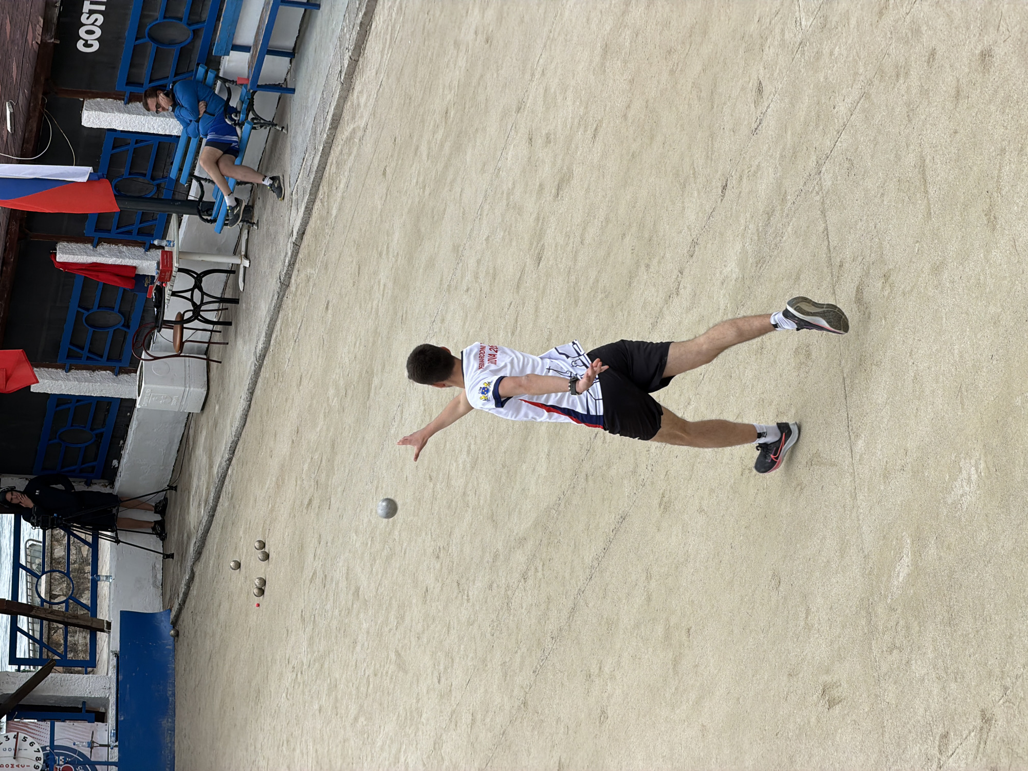 A man wearing a white and red shirt and black shorts is playing bocce in Budva, Montenegro. He is in the motion of throwing a silver bocce ball. There are several other bocce balls already on the ground near the side of the court, and a small red object. A cameraman is filming the game from behind a blue metal fence. Other people are sitting on blue benches in the background. A Russian flag hangs from the fence.