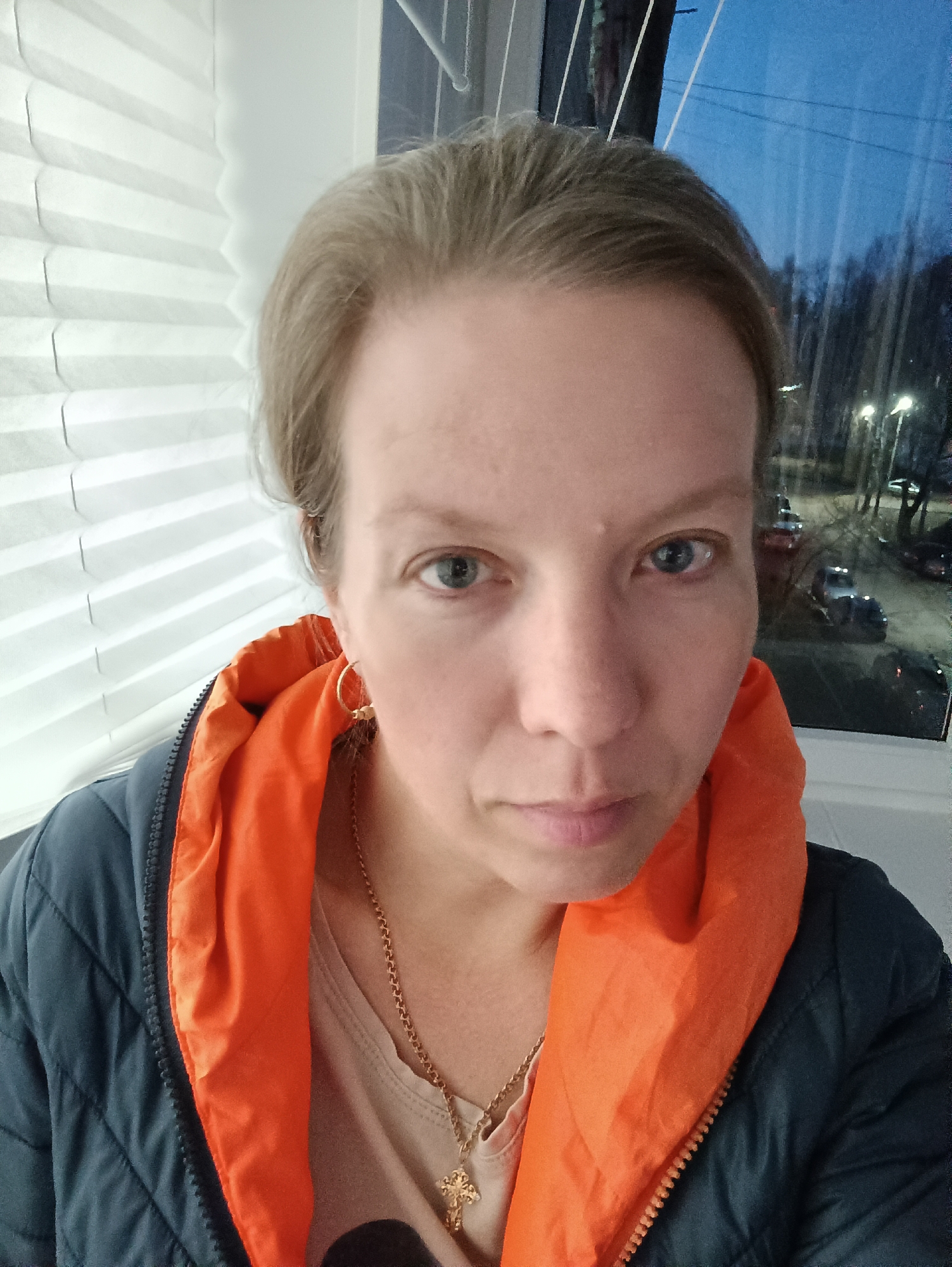 A close-up selfie captured indoors features a woman with fair skin, light blue eyes, and light brown hair pulled back from her face. She looks directly at the camera with a neutral to slightly serious expression. She is wearing a dark blue quilted jacket with a bright orange lining or hood, and a light-colored top underneath. A thin gold chain necklace with a small gold cross pendant is visible, along with a small gold hoop earring in her left ear.
To the woman's left, a window covered with white horizontal cellular blinds is visible. To her right, through another window, an outdoor scene at night or dusk is visible. The sky is dark blue, and streetlights are illuminated, casting light on parked cars and bare trees along what appears to be a residential street. The scene suggests an urban or suburban setting in Chaikovsky, Russia. There is no visible text in the image.