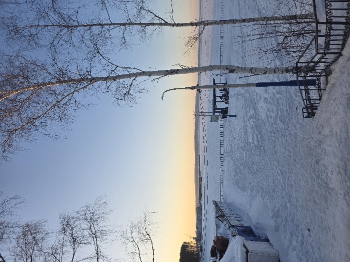 The image captures a panoramic winter scene at the frozen waterfront of the Votkinsk Reservoir in Chaikovsky, Russia, under a clear sky displaying a gradient from light blue overhead to warm orange and yellow at the horizon, indicative of sunrise or sunset.
In the immediate foreground, a snow-covered path descends to the midground, bordered on the right by a black wrought-iron railing. To the left of the path, a white, partially snow-covered structure stands adjacent to a fence topped with barbed wire, running horizontally across the scene.
The midground features an expansive, flat area covered in undisturbed snow, presumably a beach or embankment. Two prominent bare birch trees with white bark are positioned to the right of the frame, alongside a blue-colored lamppost. Further into the midground, towards the frozen water, a green-roofed, open-sided shelter is visible. Beneath this shelter, multiple individuals are discernible, accompanied by at least two snowmobiles or similar recreational vehicles, suggesting activity. Small, dark markers are sparsely arranged on the frozen surface beyond the immediate snow-covered area, forming lines.
The background consists of the vast, frozen surface of the Votkinsk Reservoir, extending to the distant horizon. The far shoreline on the left is densely forested with what appear to be coniferous trees, interspersed with industrial structures including cranes. The sky, transitioning in color, occupies the upper portion of the frame.