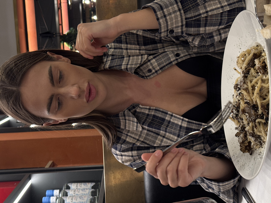 The image displays a female individual seated at a table in a restaurant or trattoria located in Verona, Italy. The woman, with long brown hair, is depicted looking downwards at a white plate of pasta, specifically spaghetti, garnished with dark truffle or mushroom pieces, grated white cheese, and black pepper. She wears a blue and white plaid long-sleeved shirt, unbuttoned to expose a black bralette or top, with a faint red mark visible on her upper chest. Her right hand holds a silver fork poised above the plate, while her left hand rests near her chin. The table is draped with a white tablecloth, displaying a silver knife and part of a wine glass to the left of the plate, and some bread pieces in a metal dish on the bottom right. The background features dark booth seating, a horizontal gold-colored surface, and an orange-toned wall. Above the gold surface, a shelf with multiple clear bottles with blue caps is visible beneath horizontal strip lighting. Blurred reflections in the distant background suggest additional interior lighting and greenery.