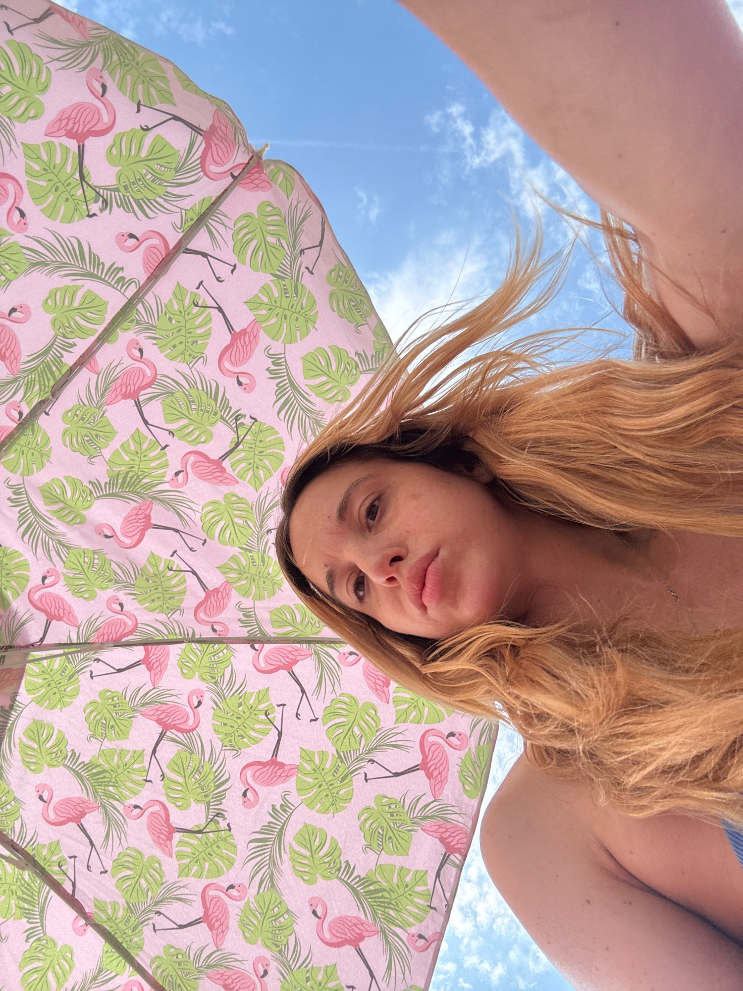 A young woman with long, blonde hair is looking up from under a pink umbrella with a flamingo and palm leaf pattern. The umbrella is partially obscuring a bright blue sky with white clouds. The woman is wearing a blue swimsuit. The image appears to have been taken outdoors, likely in a sunny location, possibly a beach or park. The perspective is from below, looking up at the woman and the umbrella. The specific location in Nesebar, Bulgaria, is not discernible from the provided image.