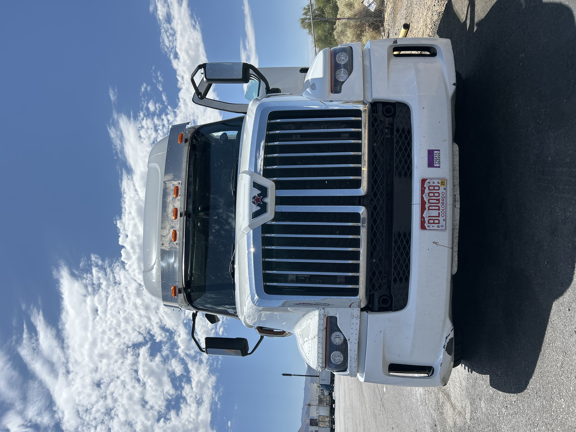 A white semi-truck is parked on an asphalt lot under a bright blue sky with scattered clouds in Primm, USA. The truck is facing the camera, showcasing its large chrome grille with a red Western Star logo. The headlights are visible, and the front bumper has some scuffs and marks. A Colorado license plate that reads "BLDQ88" is mounted on the front. A purple sticker with "NY 329535" is next to the license plate. The truck's mirrors are extended. In the background, there is a chain-link fence, dry brush, and some distant buildings. The sun casts a strong shadow of the truck onto the pavement.