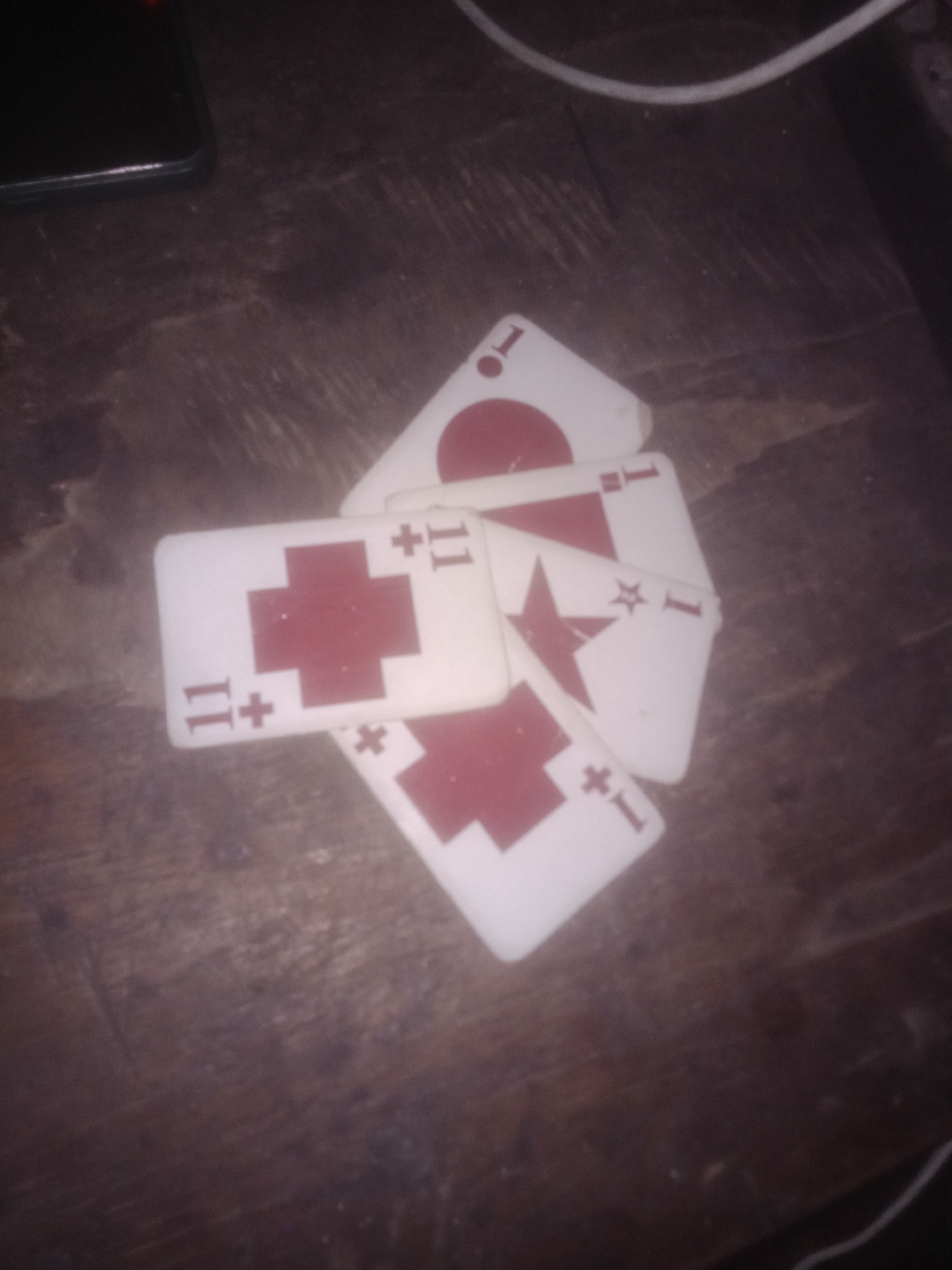 A pile of playing cards is scattered on a dark wooden surface. The cards are white with red markings. Some cards have a red cross symbol with the number "11" in each corner. Other cards have a red circle with a white "1" inside, and another card has a red star with a white "1". The lighting is dim, and the image is blurry, making it difficult to discern further details. The wooden surface appears to be a table or desk. There are no people or other objects clearly visible in the frame.