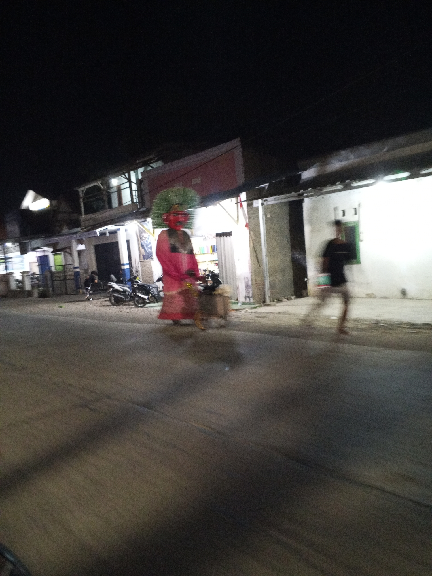 This image captures a street scene at night in Gebang, Indonesia. In the foreground, a blurry street dominates the lower half of the frame, with diagonal lines suggesting movement. To the left, a brightly lit white object, possibly a sign or light fixture, is visible. Further back, on the left side of the street, several motorcycles are parked.
The central focus of the image is a large, costumed figure, likely a "Ondel-ondel," a traditional Betawi folk art performance. The figure wears a vibrant pink garment and has a prominent green headdress and a red mask-like face. It stands near a motorcycle and a small cart. Behind the Ondel-ondel, a row of buildings lines the street. One building on the left has a balcony structure with railings. The buildings are illuminated by artificial lights, casting a bright glow on their facades.
To the right of the Ondel-ondel, a person is captured in motion, appearing as a blur as they run across the street. This individual is wearing a dark t-shirt and light-colored shorts. The overall atmosphere is one of a nighttime event or gathering, possibly a festival or celebration, given the presence of the costumed figure and the activity on the street. The image is characterized by motion blur, indicating a low light condition and a moving camera or subject.