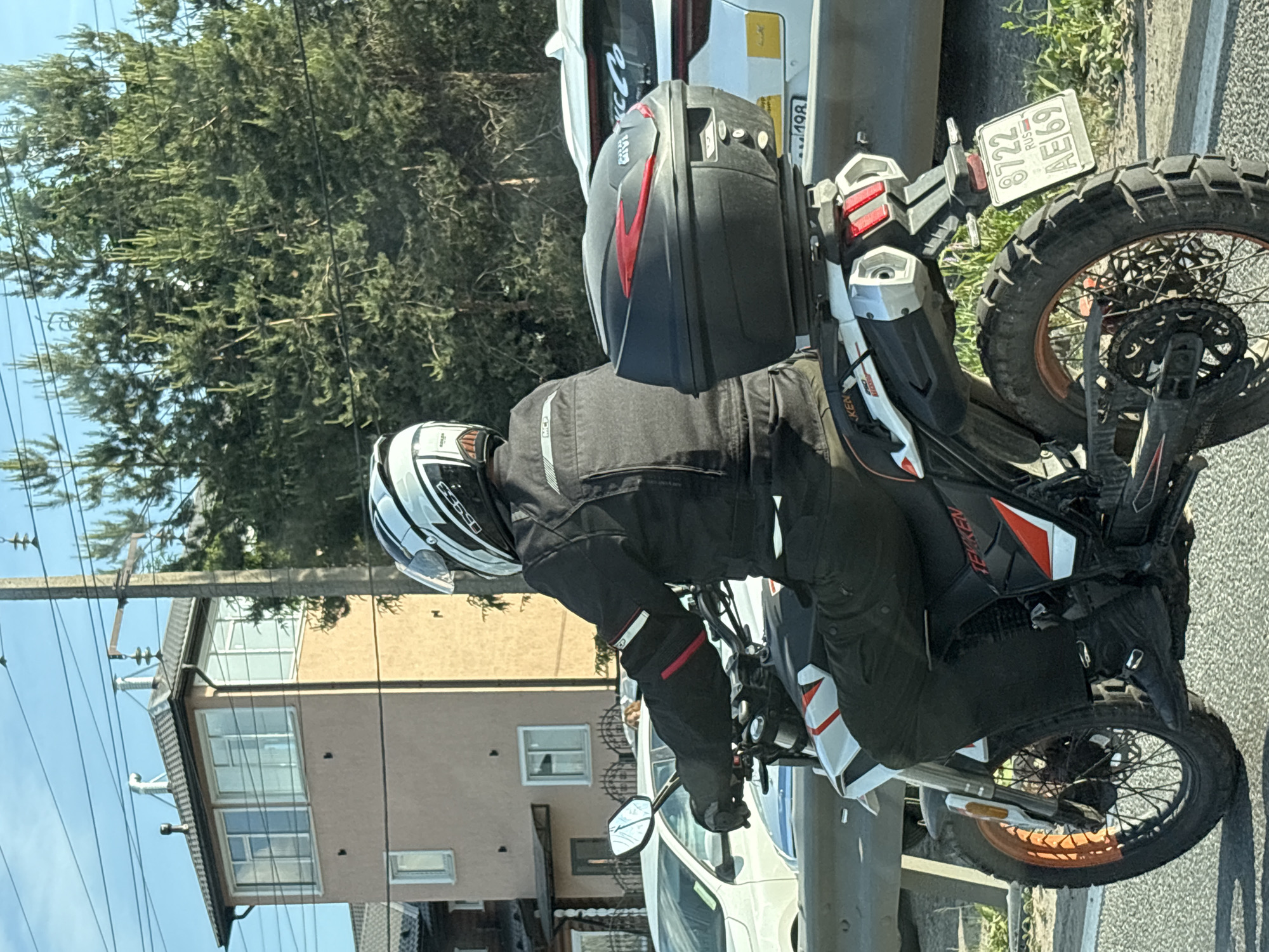 A motorcyclist is observed from behind, stationary or moving slowly on a paved road, under clear skies. The scene is set in Novosaratovka, Russia. The motorcyclist wears a white, black, and grey full-face helmet with a clear visor (brand "SOLEH" visible on the rear), a black motorcycle jacket with white and red accents (logo "MCJ" on the back), black pants, and black buckle-fastened motorcycle boots.
The individual is operating a black and white "TEKKEN" brand adventure-style motorcycle with red/orange accents. The front wheel features an orange rim and knobby tire, while the rear wheel has a black rim and a similarly knobby tire. A black "GIVI" brand top case with red accents is mounted on the rear luggage rack. The motorcycle's rear fender displays a Russian license plate: "8722 RUS AE69", with "69" indicating the Tver Oblast region.
In the background, to the left, a light brown three-story building with a dark grey hip roof is prominent. The top floor features large, white-framed windows suggesting an observation area, and two metallic ventilation pipes and a chimney are visible on the roof. Lower down, additional white-framed windows are visible. A white car, possibly a sedan, is partially visible to the left of the motorcycle, behind a metal guardrail. To the right, dense green coniferous trees are present, partially obscured by a tall wooden utility pole supporting multiple power lines. Further back, another white vehicle, likely an SUV, has a "MC Co" sticker on its rear window and "198" faintly visible on its bumper. The road surface is asphalt.