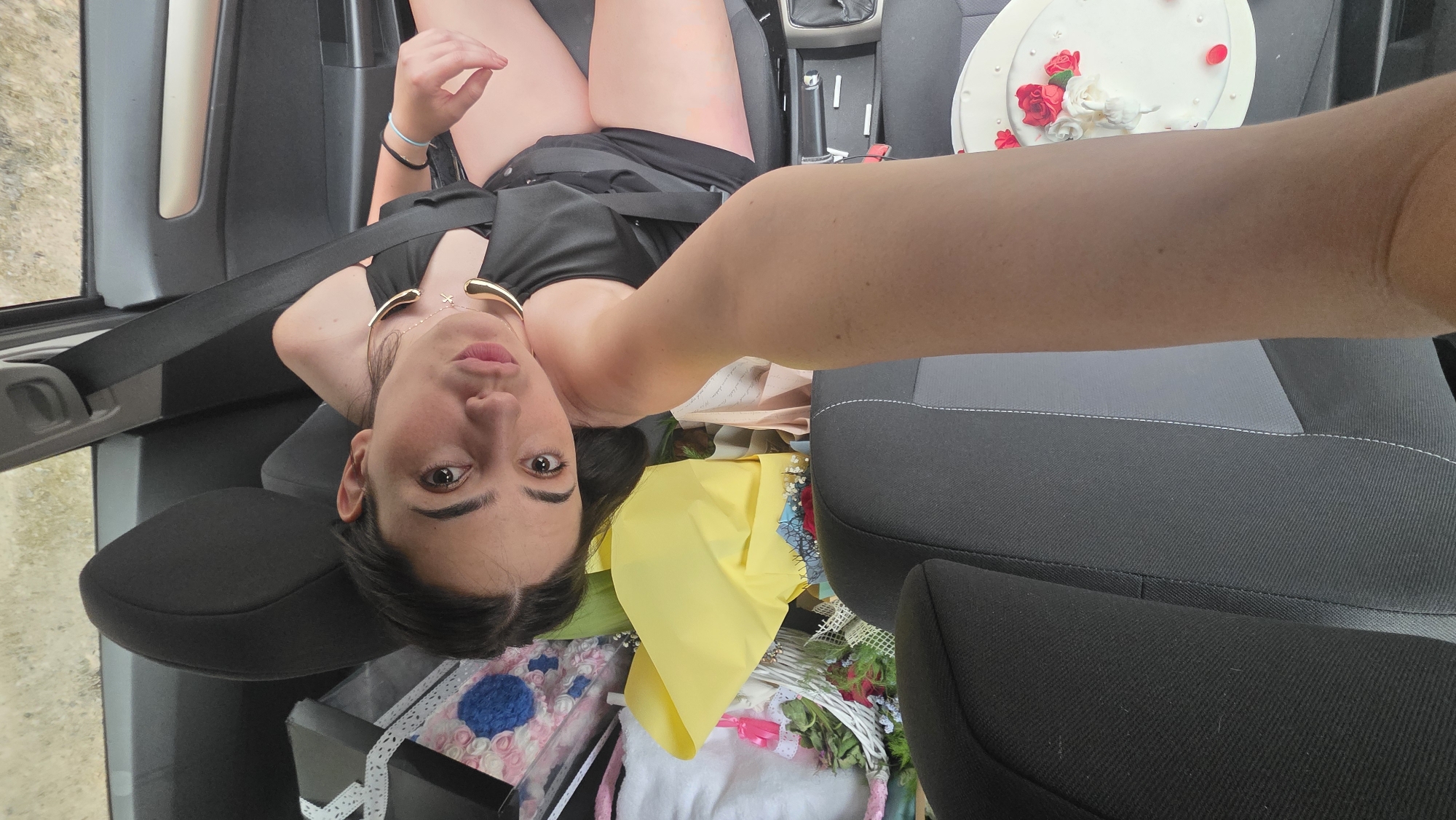 The image depicts a young woman seated in the front passenger seat of a vehicle, in Redina, Bulgaria. She is wearing a black off-the-shoulder top, a visible seatbelt across her chest, and two gold-toned necklaces, one with a star pendant and another appearing to be a choker. Her dark hair is pulled back, and she is looking directly at the camera with a slightly pouting expression. A fair-skinned human forearm, presumably belonging to the photographer, extends from the lower center of the frame, partially obscuring the front driver's seat, which is dark grey fabric with lighter grey accents and white stitching.
The area behind the woman (the backseat and floor) is filled with numerous items, including a crumpled piece of bright yellow fabric, a white woven basket containing red roses and other greenery, a blue and pink floral-patterned item, a white fluffy object, a black gift box secured with a white ribbon, and a light brown paper bag. On the passenger side floor, near the center console, a white circular cake decorated with red and white roses and scattered red petals is visible. The exterior view through the car window suggests an outdoor, possibly unpaved or dirt road environment, with blurry background elements.