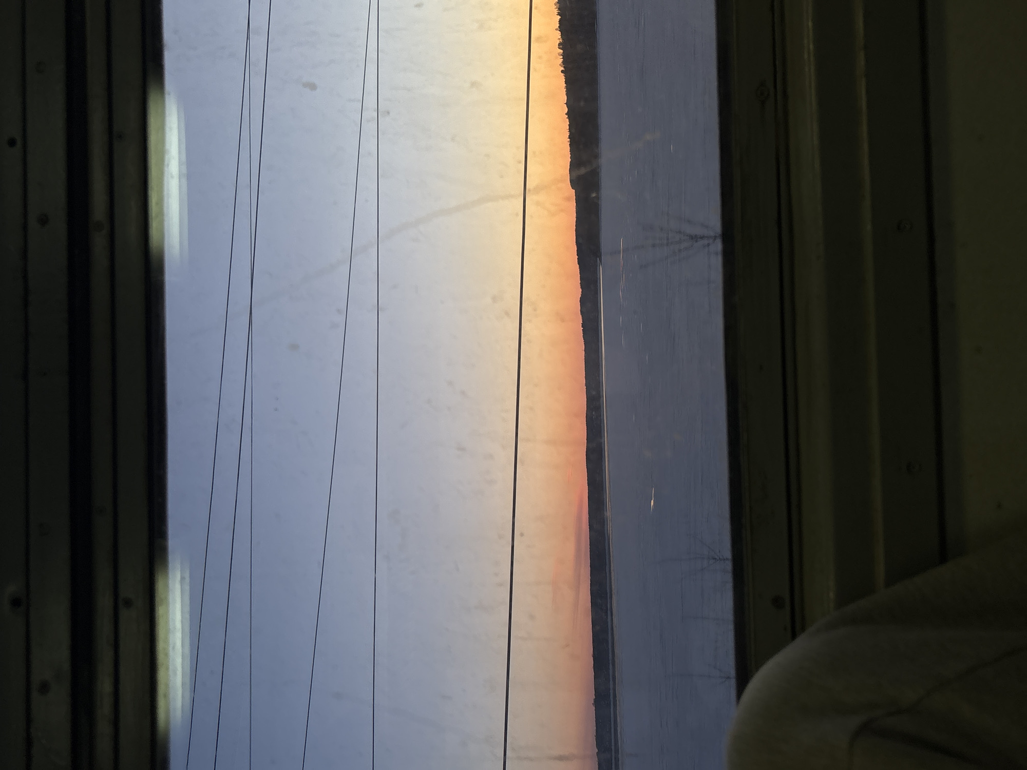 A view from a train window shows a landscape at sunset. Power lines run vertically in the left side of the frame, obscuring a view of the sky. A body of water is visible to the right of a strip of land with silhouetted trees, all reflecting an orange sunset. The window frame is visible on both sides. Part of the train interior is visible in the bottom right. This photograph was taken in Пальники, Russia.