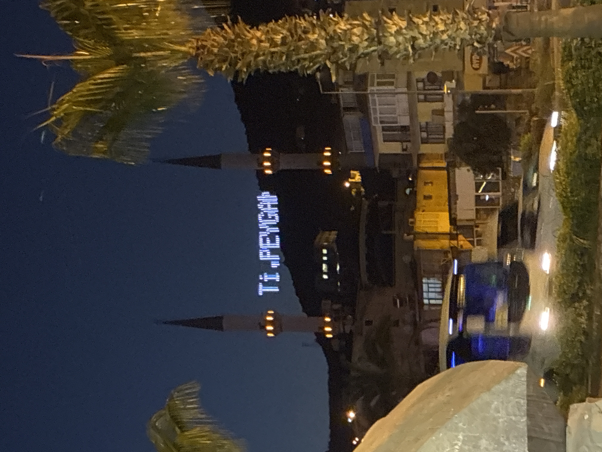 The image captures a nighttime view of a mosque in Alanya, Turkey. Two minarets flank the central structure of the mosque, with bright, white LED lights illuminating the words "Tİ.PEYCA" on the hillside behind it. Below the mosque, a street level view shows a busy road with cars and a bus in motion, their headlights creating streaks of light. Buildings and palm trees are also visible, illuminated by various lights, contributing to the vibrant nighttime urban landscape of Alanya.