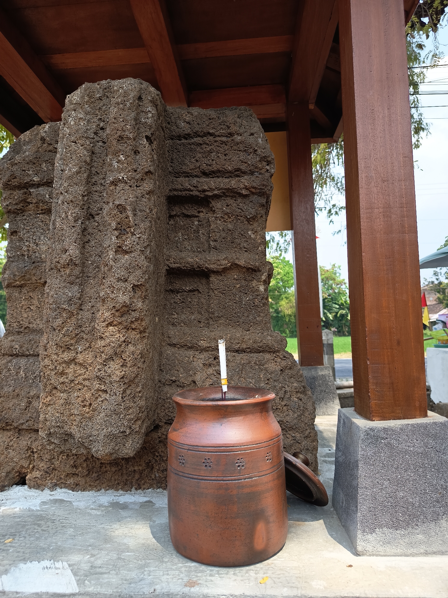 The image shows a terracotta jar with a lit cigarette in it, placed on a concrete surface in front of a large, rough-hewn stone structure. The stone structure appears to be part of an ancient ruin or monument. Behind the stone, a wooden roof structure supported by thick wooden pillars is visible. Green trees and a bright sky are visible in the background, suggesting an outdoor setting. The location is identified as the city of Lalung, Indonesia. The jar is decorated with horizontal bands and small floral patterns. The lid of the jar is placed beside it. The stone structure has a rough, porous texture with vertical indentations. The overall scene depicts an outdoor area with historical elements and everyday objects.