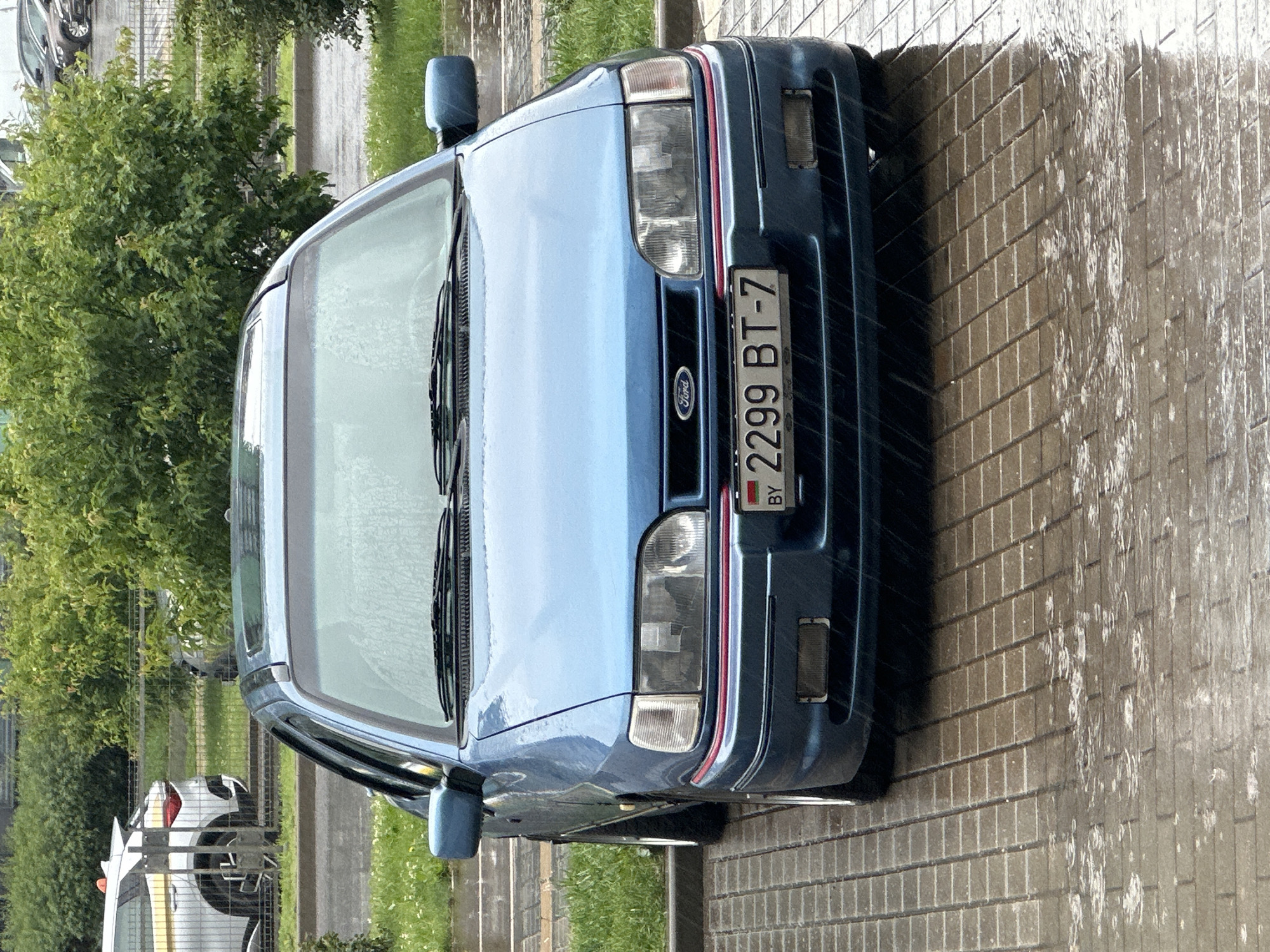The image captures a blue Ford Sierra parked on a wet, tiled surface in what appears to be an outdoor setting. Rain is visible falling, indicated by streaks on the car and puddles on the ground. The car is the primary subject, with its front prominently displayed. The license plate reads "2299 BT-7" with "BY" in the corner, suggesting it is registered in Belarus. In the background, there are green trees and bushes, as well as a glimpse of other vehicles and a fence. The overall impression is of a car parked in a city during a rain shower.