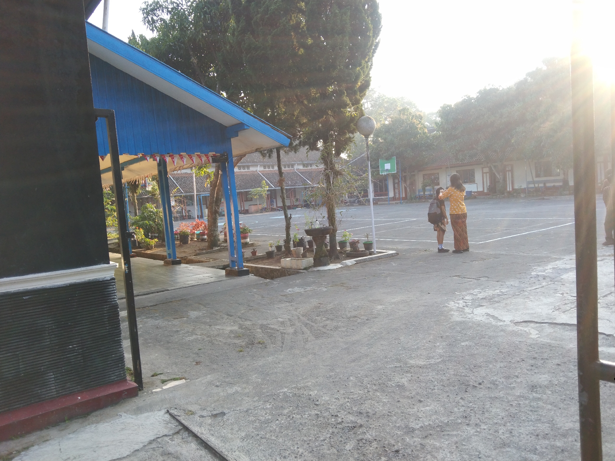 The image shows an outdoor scene at what appears to be a school or community area in Magelang, Indonesia. In the foreground on the left is a dark, textured wall with a white trim and a reddish-brown base, possibly part of a building. Adjacent to this wall is a black vertical pole. To the right of the pole is a blue building with a white-tiled roof and some red and white decorative flags hanging from the eaves. Beyond the blue building, the ground is paved with gray concrete.
In the mid-ground, two people are standing on a gray paved court, which might be a basketball court given the presence of a green basketball hoop in the background. One person, wearing a yellow patterned top and a patterned skirt, stands facing away from the camera, interacting with another person. This second person, appearing to be younger, is also facing away and is wearing a backpack. The paved area has white lines marking boundaries.
The background features several buildings with white walls and tiled roofs, typical of Indonesian architecture, and numerous trees with dense foliage. The sun is shining brightly, creating lens flare on the right side of the image and casting a warm, bright light on the scene, suggesting it is either early morning or late afternoon. The environment appears to be a schoolyard or an open public space with recreational facilities.