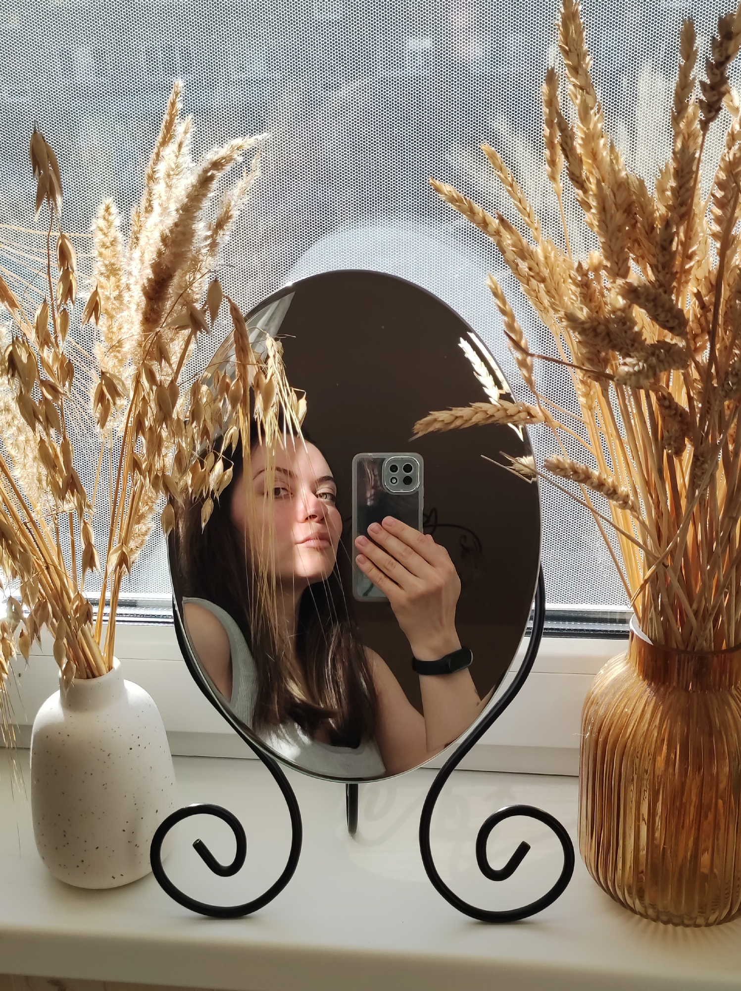 A woman is taking a selfie in a mirror that is placed on a windowsill. The mirror is oval-shaped with a black metal stand featuring curled ends. The windowsill is white and appears to be behind a curtain. On either side of the mirror are vases: a white vase on the left and an amber-colored vase on the right. Both vases contain dried wheat or grass arrangements. The woman is holding a smartphone with a clear case and multiple camera lenses. She is wearing a black wristband. The location is Chaikovsky, Russia.