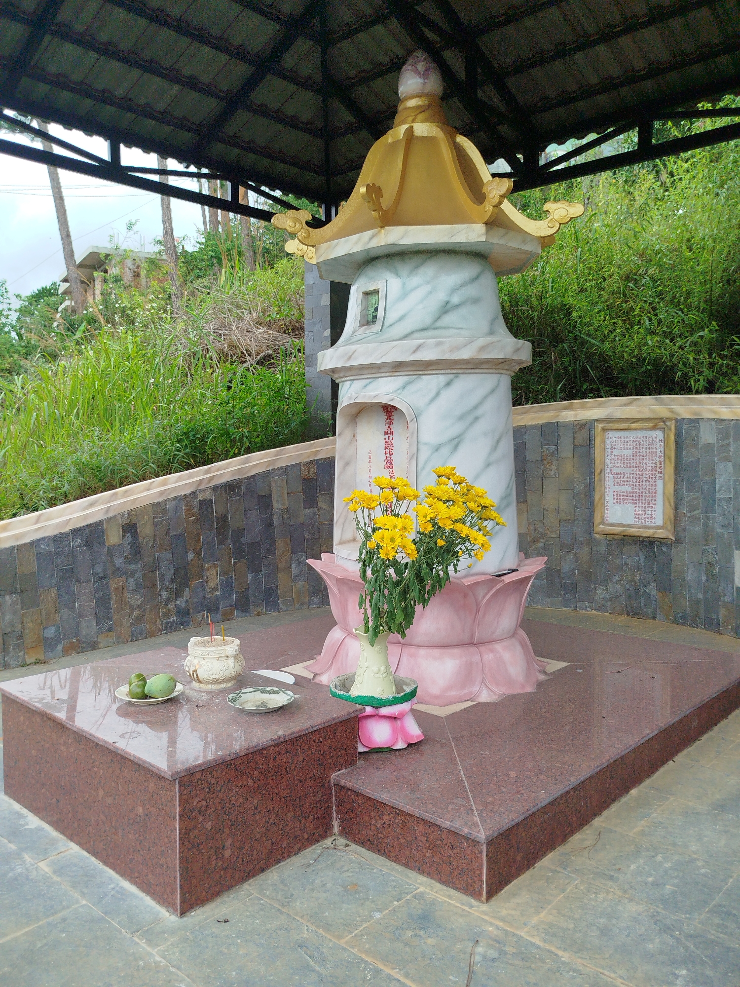 This image depicts an outdoor shrine located in Da Lat, Vietnam. The shrine is situated under a covered structure with a dark, tiled roof supported by dark metal beams. At the center of the scene is a multi-tiered monument, with a tall, cylindrical central section adorned with a pale, marble-like pattern. This central section is topped with a decorative gold finial. The base of the monument is designed to resemble pink lotus petals.
In front of the monument, a bouquet of bright yellow chrysanthemums is displayed in a vase. To the left of the monument, on a raised, polished red granite platform, are several offerings: a small white ceramic incense burner with three incense sticks, two green fruit, possibly limes or small mangoes, on a white plate, and a small dish with a light-colored substance.
The monument is surrounded by a polished red granite base and walkway, with darker stone tiles forming the ground surface. Behind the shrine, a textured stone wall rises, covered in various shades of gray and brown rectangular stones. Beyond the wall, lush green vegetation and trees cover a hillside. To the right of the main monument, a framed plaque with red Chinese characters is affixed to the stone wall. The overall setting suggests a place of remembrance or worship, possibly a family grave or a memorial, in a natural, elevated environment characteristic of Da Lat. The sky is overcast, suggesting a cloudy day. No people are present in the image.