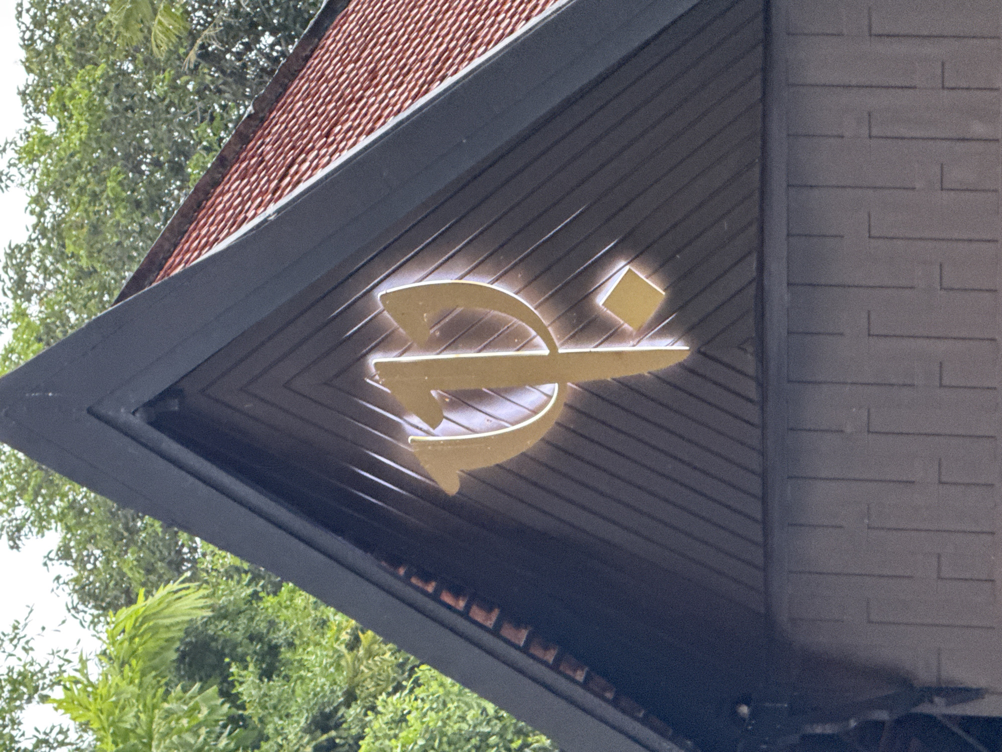 The image displays a close-up, eye-level shot of a building's architectural detail. The primary subject is a large, golden, stylized emblem mounted on a dark, angled wall paneling. The emblem has a flowing, calligraphic appearance, with a curve and a square element. It is illuminated from behind, creating a glowing effect. Above the emblem, a portion of a red, tiled roof is visible. To the left of the building's facade, lush green foliage from trees and plants is seen against a bright sky, suggesting an outdoor setting during the daytime. The building's facade is constructed with dark, horizontal planks, and to the right, a textured gray wall with a brick-like pattern is visible. The scene suggests the entrance or exterior of a building, possibly a hotel, resort, or establishment with a distinct brand identity. The lighting and clear visibility of the emblem indicate it is daytime, and the foliage suggests a tropical or sub-tropical environment. No people are visible. There is no visible text. The location context is Karon, Thailand.