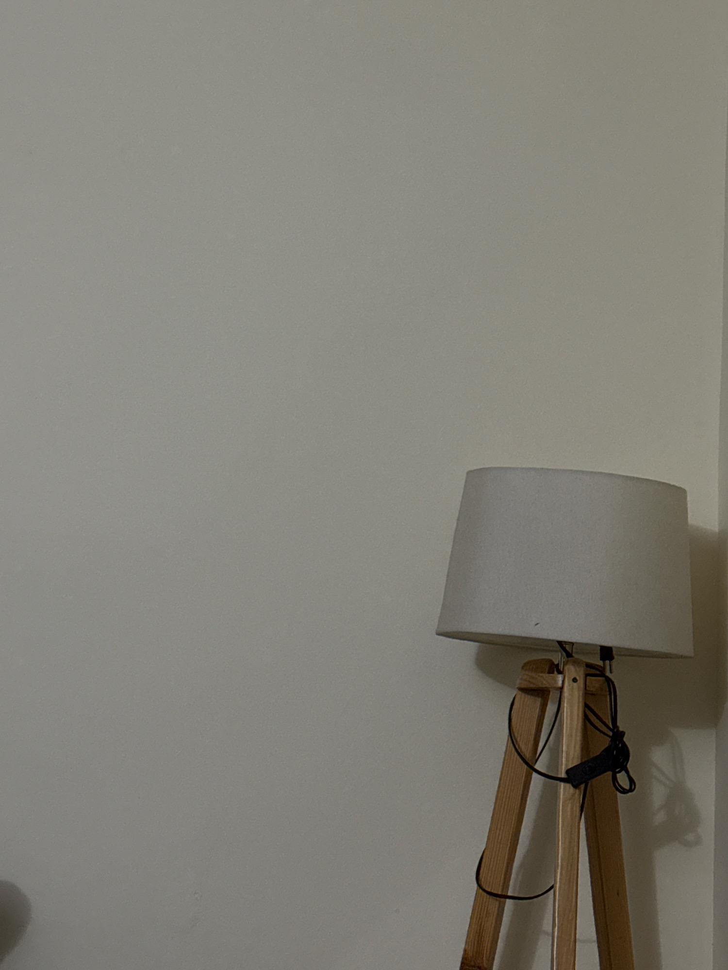 The image depicts a floor lamp with a light-colored fabric shade and a wooden tripod base. A black power cord is visible, wrapped around one of the legs of the tripod and secured with a black clip. The lamp is positioned in a corner, casting a shadow on the wall. The background is a plain, light-colored wall. The image quality suggests it was taken with a mobile device. No specific location in Kohistan Enclave, Pakistan, is indicated by the visual content.