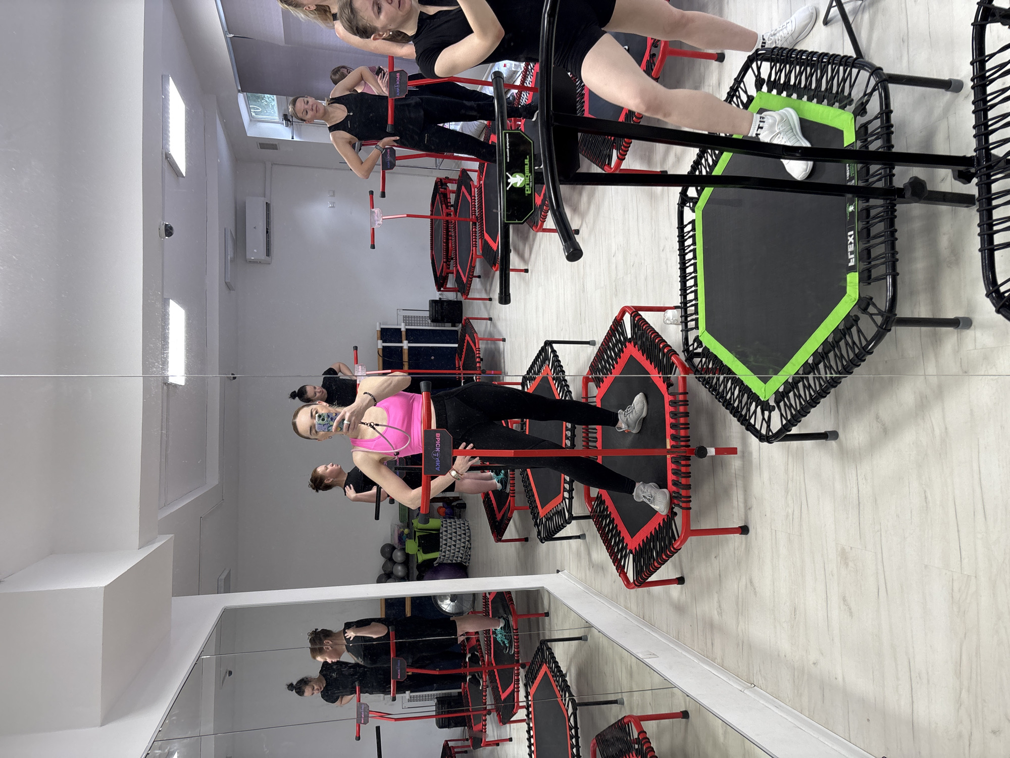 The image shows a group of women in a fitness studio, likely a gym, participating in a trampoline fitness class. The room is brightly lit and features a large mirror along one wall, reflecting the scene. Several mini-trampolines, mostly red and black with some green accents, are arranged throughout the space.
In the foreground, a woman in a bright pink sports bra and black leggings is taking a selfie in the mirror, holding her phone. She is standing on a red and black trampoline. To her right, another woman in a black tank top and black shorts is visible, also on a trampoline. Further back, several other women are seen on trampolines, some in black athletic wear. The studio appears to be equipped with modern amenities, including air conditioning and lighting systems, visible on the ceiling. The floor is light-colored and appears to be wood or laminate. The overall impression is a dynamic and active fitness environment. The location is identified as Unknown, Russia.