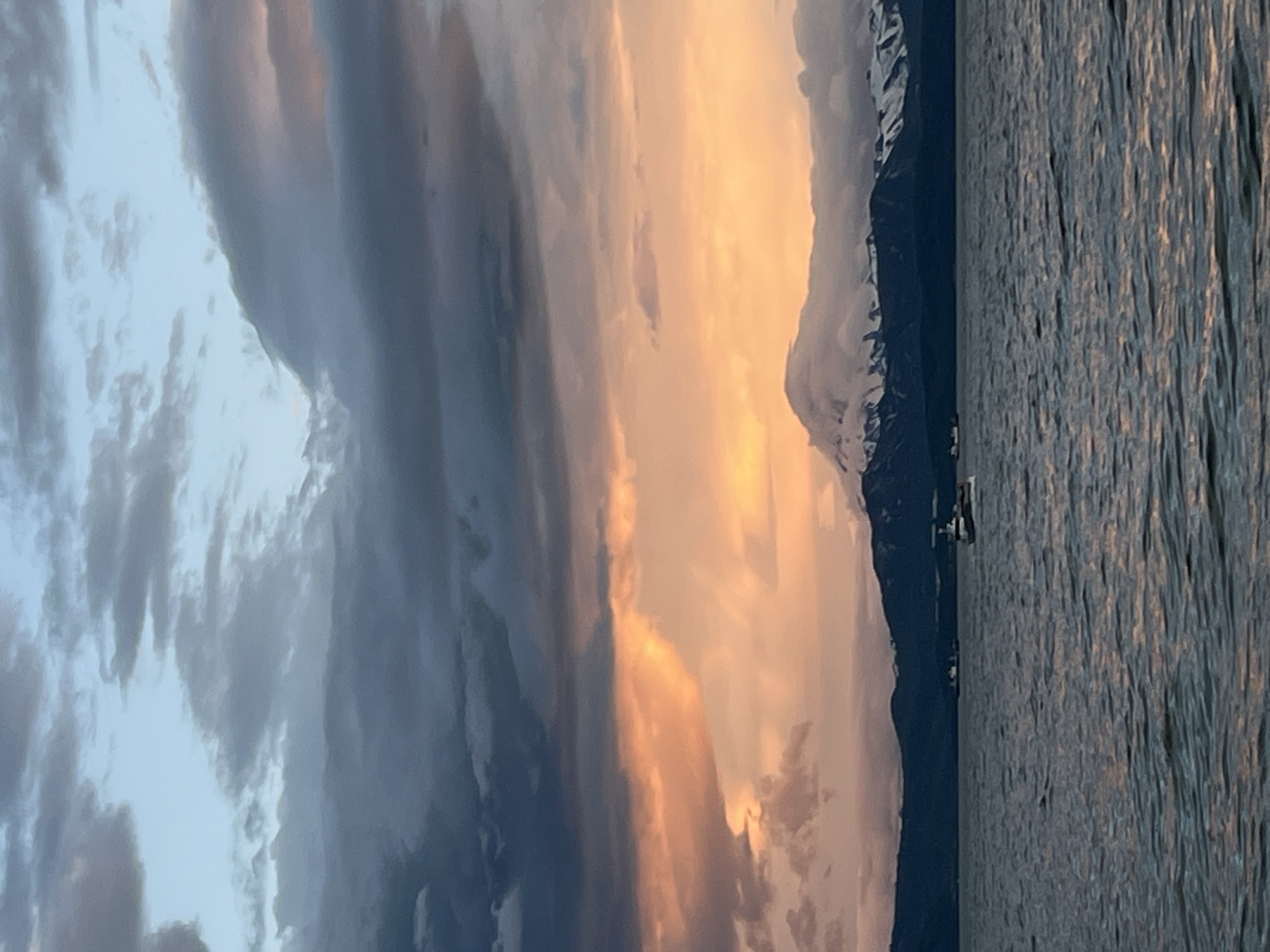 This is a medium-shot, vertical, outdoor, daytime photograph capturing a dramatic sunset over a body of water, with a snow-capped mountain range in the background. The sky is filled with dark, heavy clouds tinged with vibrant orange and pink hues from the setting sun. A few lighter blue patches are visible at the top. In the middle ground, a dark, silhouetted mountain range is visible, with its peaks covered in snow and partially obscured by clouds. A large, dominant mountain with a snow-covered summit stands out prominently. The foreground is dominated by a vast expanse of dark, choppy water, with subtle reflections of the sky. Two small boats are visible on the water, one closer to the right side of the frame and another further to the left. The scene appears to be a natural landscape, possibly near Yelizovo, Russia, given the typical volcanic and coastal geography of the Kamchatka Peninsula.