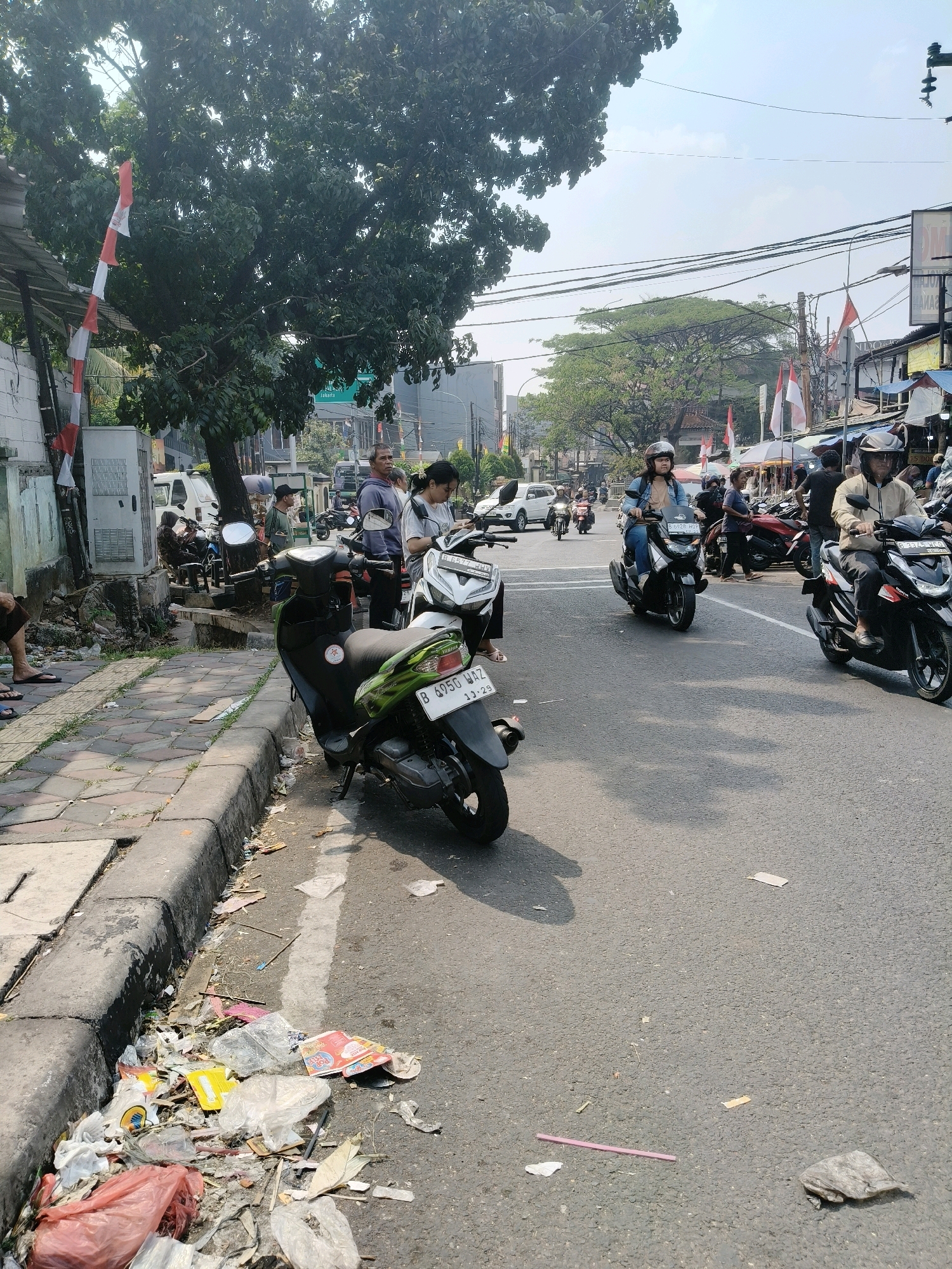 The image captures a street scene in South Tangerang, Indonesia, on a bright, sunny day. The foreground is dominated by a pile of garbage collected along the curb, consisting of plastic bags, food wrappers, and other debris. A green and black scooter is parked on the street, partially obscuring the curb. A sidewalk made of interlocking paving stones runs alongside the garbage.
Further down the street, several people are present, some on motorcycles and others standing. A woman in a blue jacket rides a black scooter, wearing a helmet. Another person in a light-colored jacket rides a black scooter, also with a helmet. In the background, more motorcycles are visible, as well as buildings with awnings and signage. Red and white Indonesian flags are displayed on poles along the street. The sky is clear, with some power lines visible overhead. The overall impression is of a busy urban environment with a notable amount of litter.