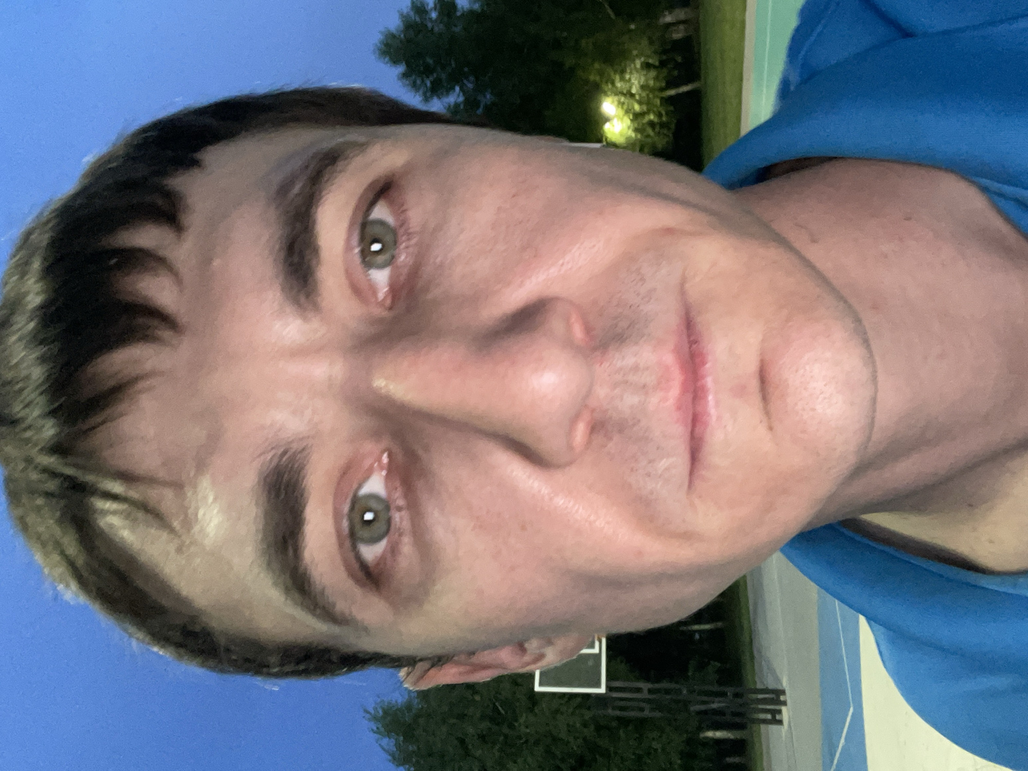 A close-up, full-face, outdoor, selfie-style photograph captures a Caucasian male with light skin and dark brown hair. The man's eyes are open and looking directly at the camera, and his mouth is closed in a neutral expression. He is wearing a bright blue polo shirt. The background suggests an outdoor recreational area, possibly a basketball court, with trees and lighting visible behind him. The sky is a deep twilight blue, indicating it is either early morning or late evening. The location is identified as Laishevo, Russia.