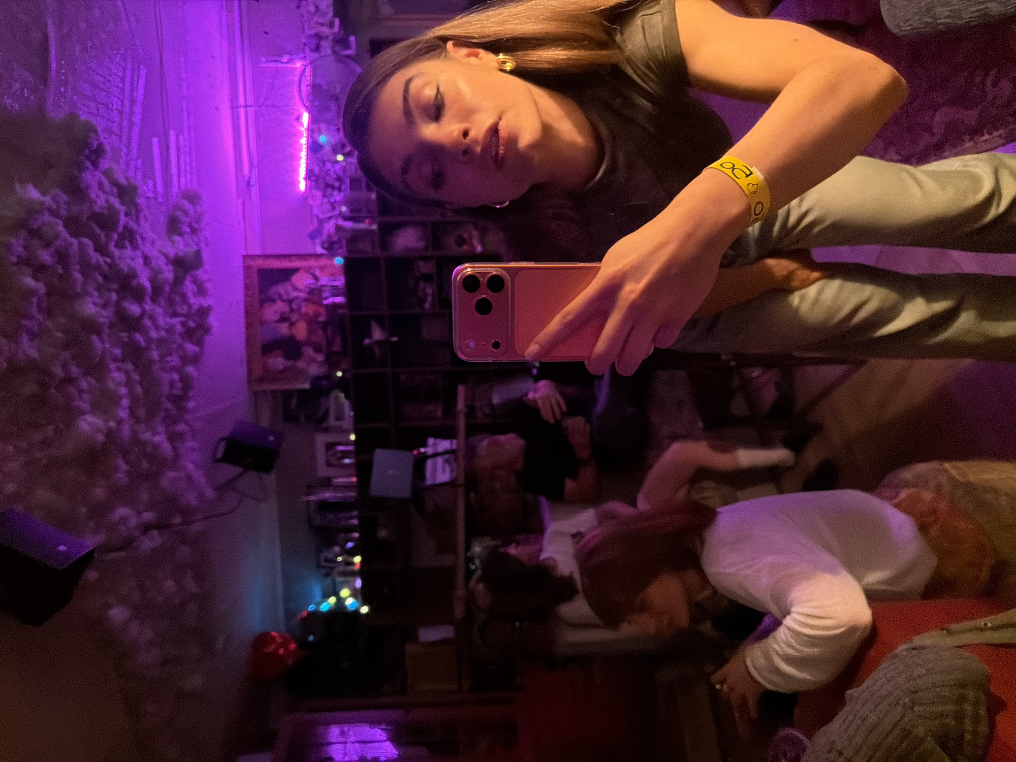 The image is a selfie taken by a young woman in a vibrant, dimly lit indoor setting, heavily saturated with purple light. The original photograph is rotated 90 degrees clockwise.
The main subject, a woman with long, straight brown hair, gazes directly into the camera with a poised expression. She is dressed in a dark green top and light-colored pants. On her right wrist, she wears a yellow wristband featuring "20" and small heart shapes. She holds a light pink/peach colored smartphone, identifiable by its triple-lens camera system, to capture the image. She has prominent gold hoop earrings.
In the blurred background, other individuals are partially visible, suggesting a social gathering. One person wearing a white long-sleeved shirt and another with light-colored hair are discernible.
The setting is an indoor space, likely a club, bar, or event venue, understood to be in Moscow, Russia. The decor is artistic and moody, characterized by a wall or art installation covered in a fluffy, cloud-like material on the left, intensely illuminated by purple light. A framed picture or painting with colorful abstract elements hangs on a wall. A dark shelving unit holds various indistinct items. String lights with blue, green, and red bulbs contribute to the festive ambiance. Small speakers and a red decorative object are also visible towards the lower part of the scene.
The artificial lighting confirms the time of day as evening or night. The overall atmosphere is lively and social.