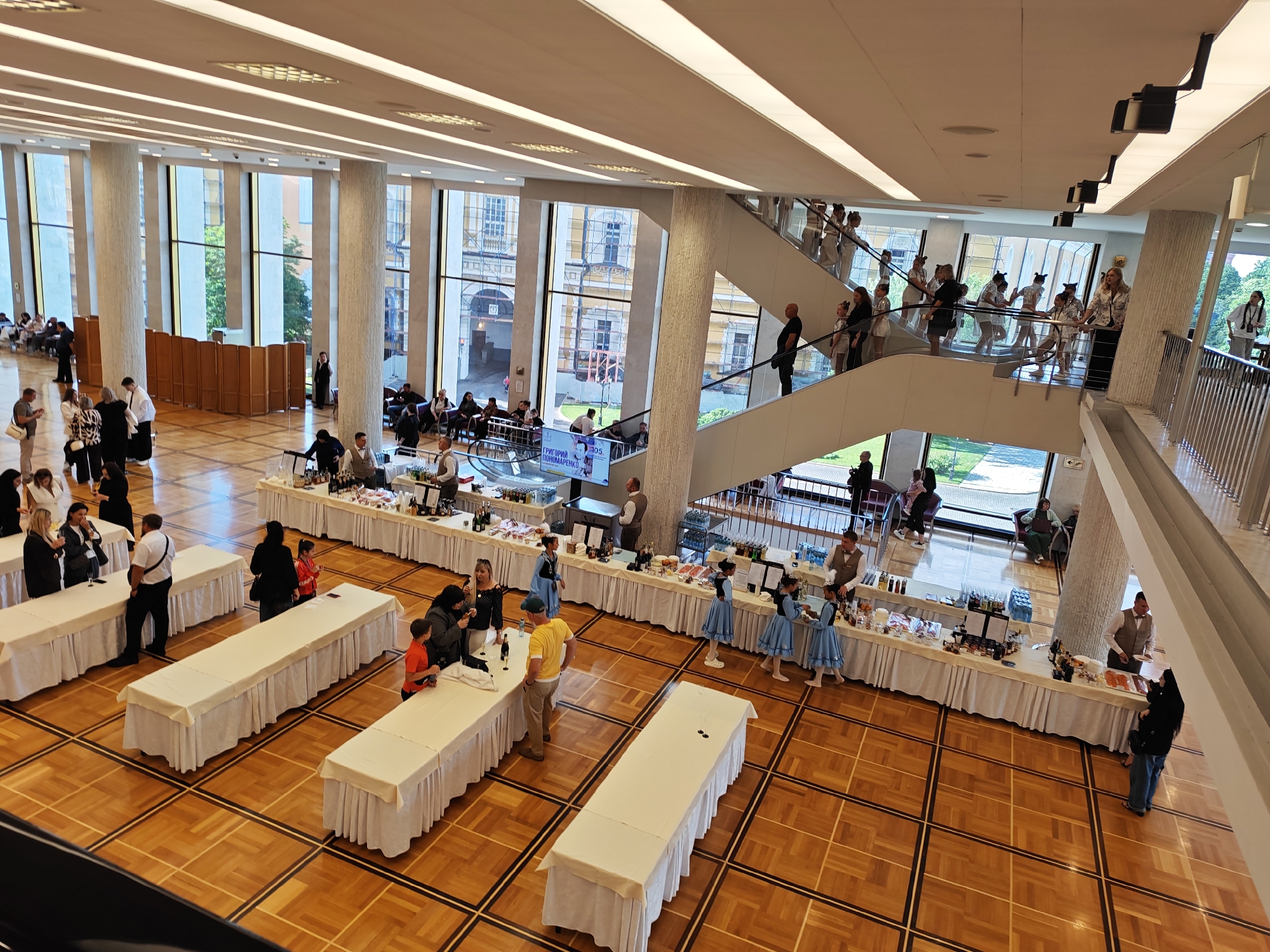 This is a high-angle, wide shot of a large, well-lit indoor event space with polished wooden floors. The space appears to be a reception or exhibition hall, featuring numerous long tables draped with white tablecloths, some laden with food and drinks. In the foreground, a few people are gathered around the tables.
Further into the hall, a tiered seating area is visible on the left, with individuals seated. On the right, a prominent staircase with a glass railing ascends to an upper level. Several young girls in blue dresses are seen on the staircase and the upper level, suggesting a performance or special event. Adults are also present on the staircase and mingling in the background, some appearing to be staff. The architecture includes large windows that allow natural light to enter, and the ceiling is modern with integrated lighting. The scene conveys a sense of organized activity and public gathering. There is no indication of the location being in Brekhovo, Russia, or any specific city.