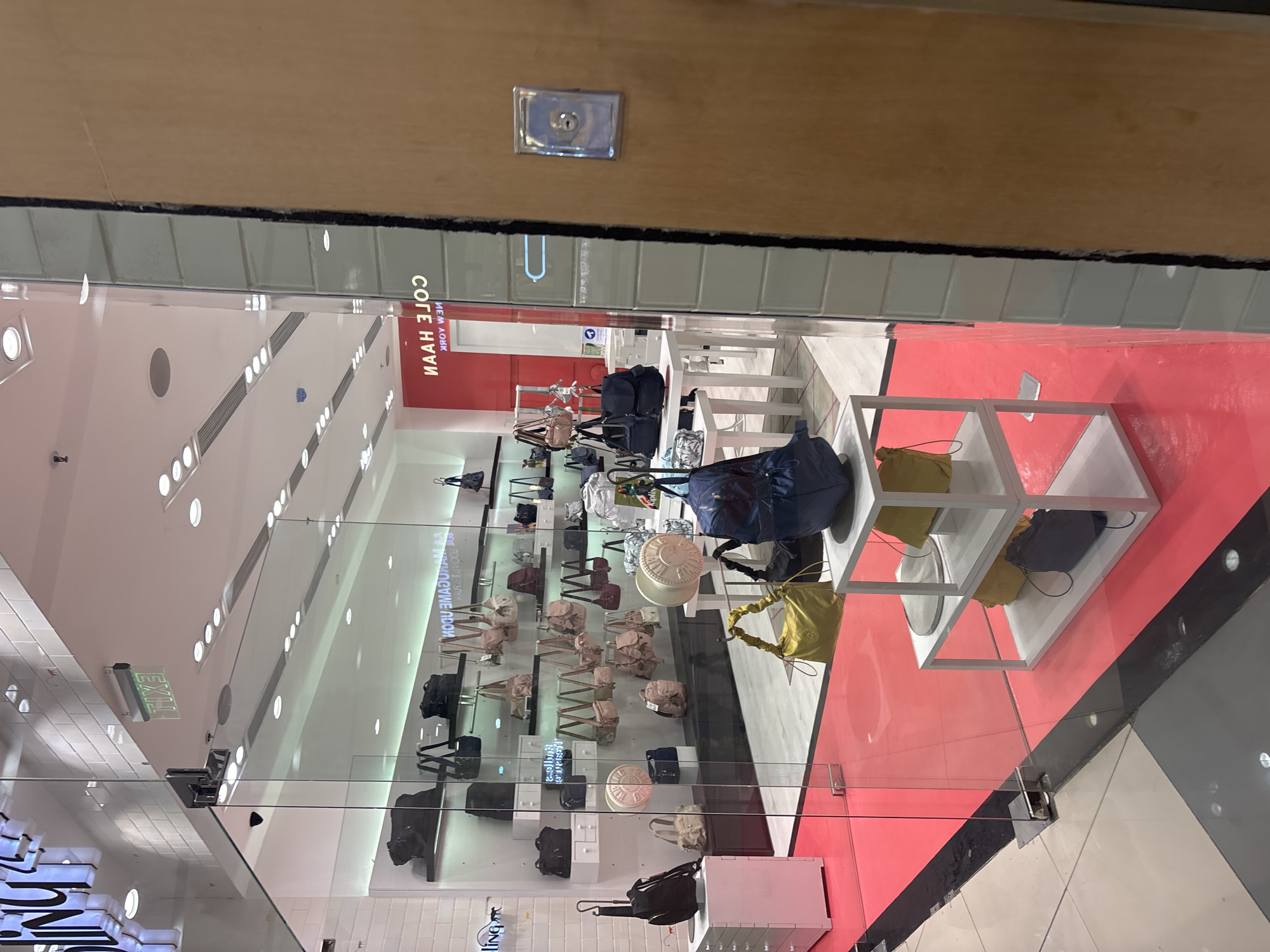 The image captures the interior of a retail store, specifically a Kipling store located in Davao City, Philippines, as indicated by the reflective signage visible in the background that reads "COLE HAAN NEW YORK" and the presence of Kipling bags, a brand commonly found in department stores and dedicated boutiques. The store features a modern display with shelves showcasing a variety of handbags in different colors and styles. The flooring is a combination of red and dark gray tiles, with a red carpeted walkway leading into the store. In the foreground, several bags are displayed on white shelving units, including a prominent yellow handbag and a large blue backpack. The store's interior is well-lit, with bright overhead lighting illuminating the merchandise. The exterior is characterized by a glass storefront and doors, allowing a clear view of the products inside. A security camera is visible mounted on the ceiling near the entrance.
