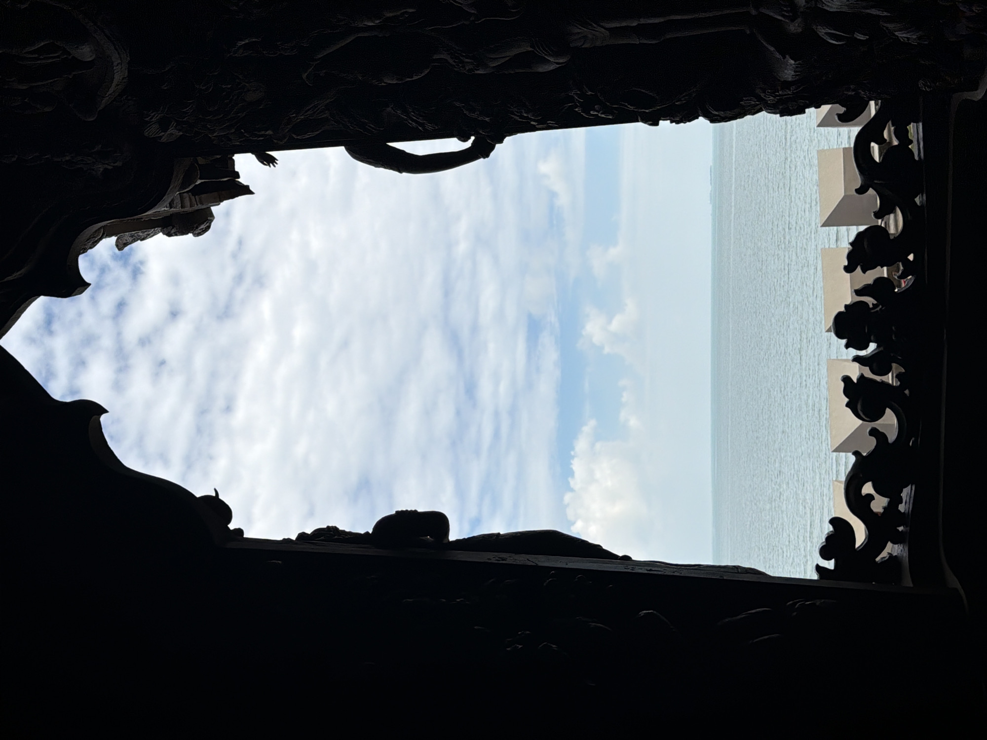 This image captures a view of the sea and sky through a intricately carved wooden archway. The archway is dark and silhouetted, obscuring much of the detail of its carvings, which appear to depict figures and ornate patterns. Through the opening, a calm sea with gentle waves stretches to the horizon, where a faint outline of land or a ship can be seen. The sky above is a mix of pale blue and white clouds, with the light suggesting it is daytime. In the foreground, a decorative wooden railing with a swirling, organic design runs along the bottom of the archway. The overall impression is one of peaceful contemplation of nature framed by artistic craftsmanship. This scene is characteristic of the Sanctuary of Truth in Pattaya City, Thailand, a magnificent wooden structure adorned with intricate carvings that overlooks the sea.