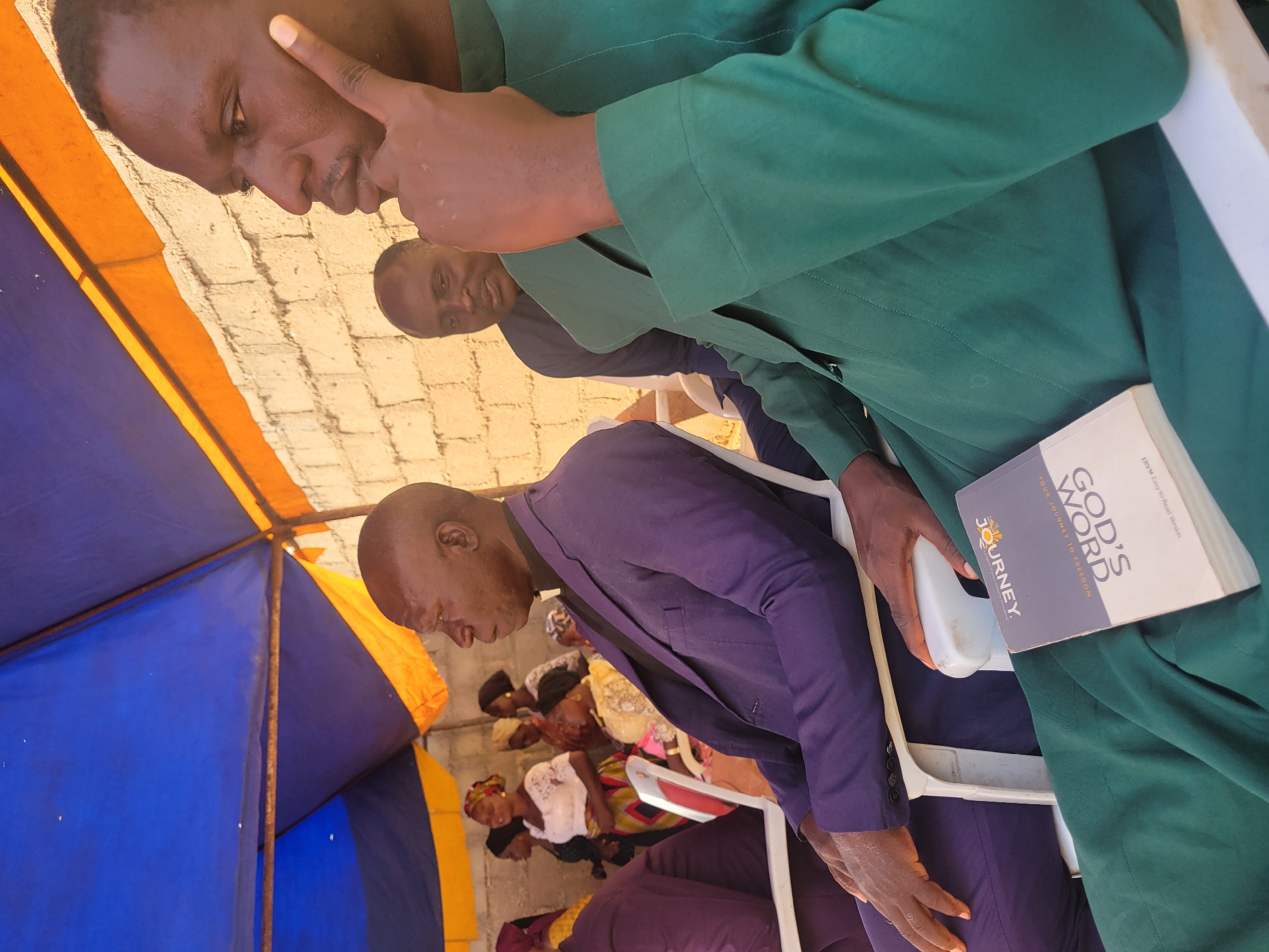 In Madalla, Nigeria, under a blue and orange tent, a group of people are gathered. A man in a purple suit and a white clerical collar sits in the foreground, facing left. To his right, another man in a green robe is seated, holding a book titled "GOD'S WORD YOUR JOURNEY TO FREEDOM." In the background, other men and women are visible, some standing and others seated. The setting appears to be outdoors, with a brick wall behind the tent. The overall atmosphere suggests a religious gathering or service.