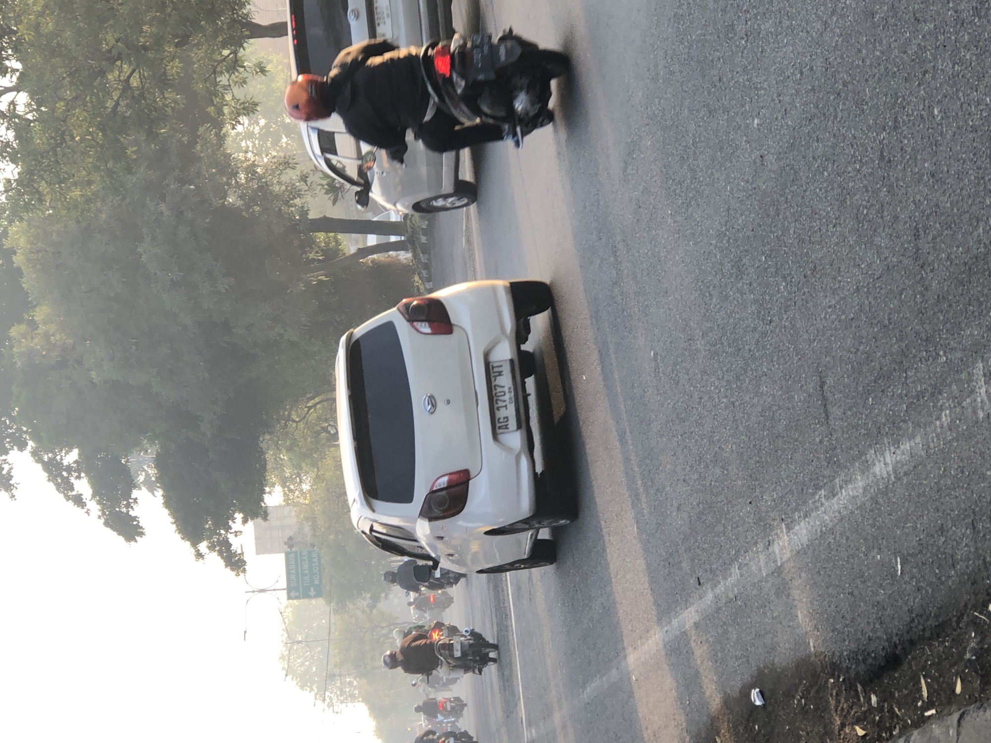 In Kludan, Indonesia, on a hazy morning, traffic flows along a paved road. A white Daihatsu Agya, license plate AG 1707 WT, is in the foreground, seen from the rear three-quarters angle. To its right, a motorcyclist wearing a red helmet and dark clothing rides a dark-colored scooter, also in motion. Behind the Agya, a line of motorcycles and riders stretches into the distance, with some vehicles blurred by motion. To the far right, a white car, partially visible, is also in motion. Overhead, a directional sign points towards Surabaya, Tulangan, and Mojosari. The environment is characterized by trees with dense foliage and a bright, hazy sky, suggesting a potentially polluted or humid atmosphere. The road surface is asphalt, marked with white lines. Some litter, including a crumpled white object, is visible on the roadside.