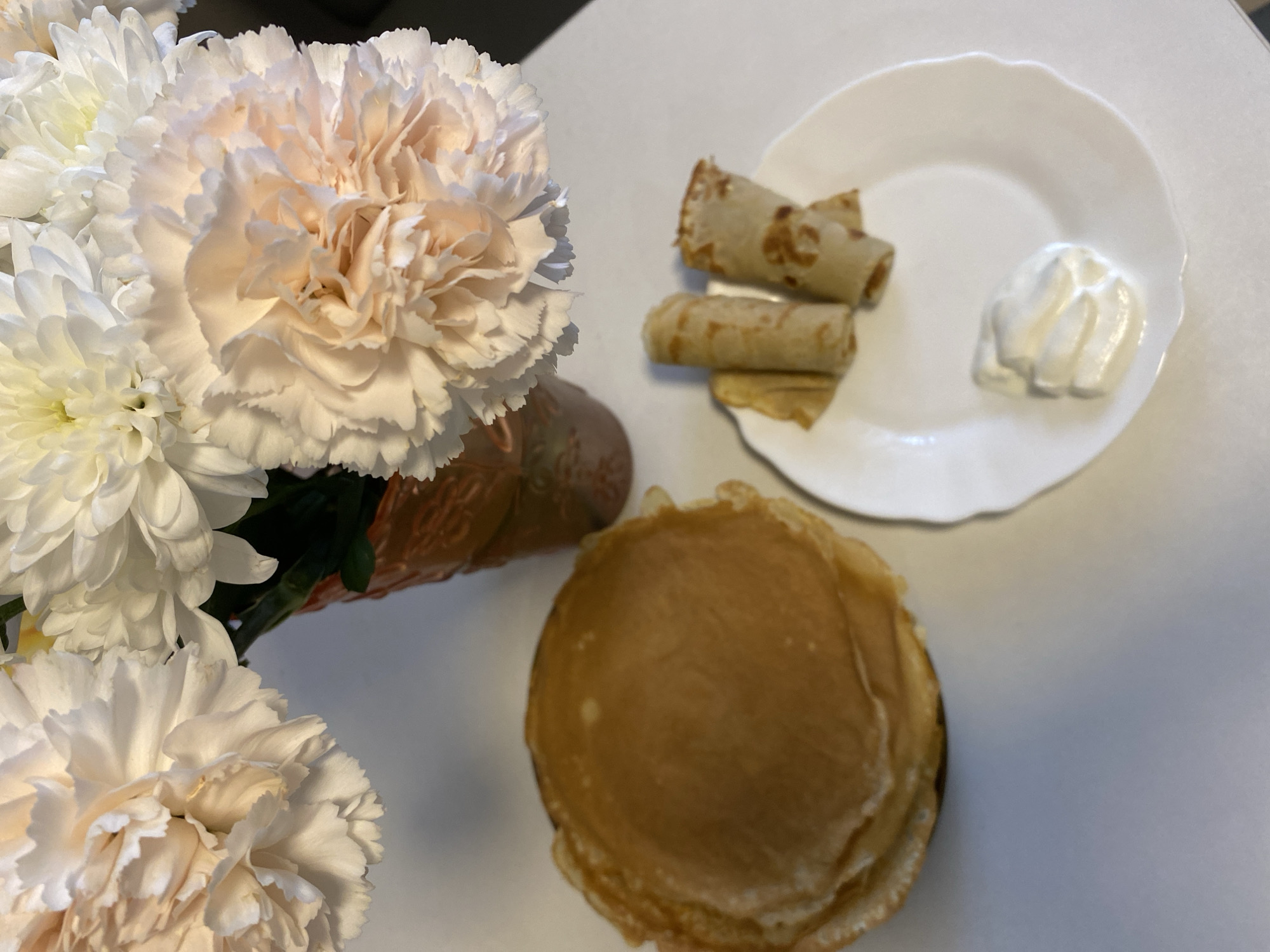 The image depicts a tabletop still life. On the left, a bouquet of white flowers, possibly chrysanthemums and carnations, sits in a patterned vase. To the right, a white plate holds two rolled crepes or blini, and a dollop of white cream, possibly sour cream. Below the plate is a stack of unfolded pancakes or blini. The background is a neutral white. The city or region of Noviy, Russia, is not identifiable from the scene.