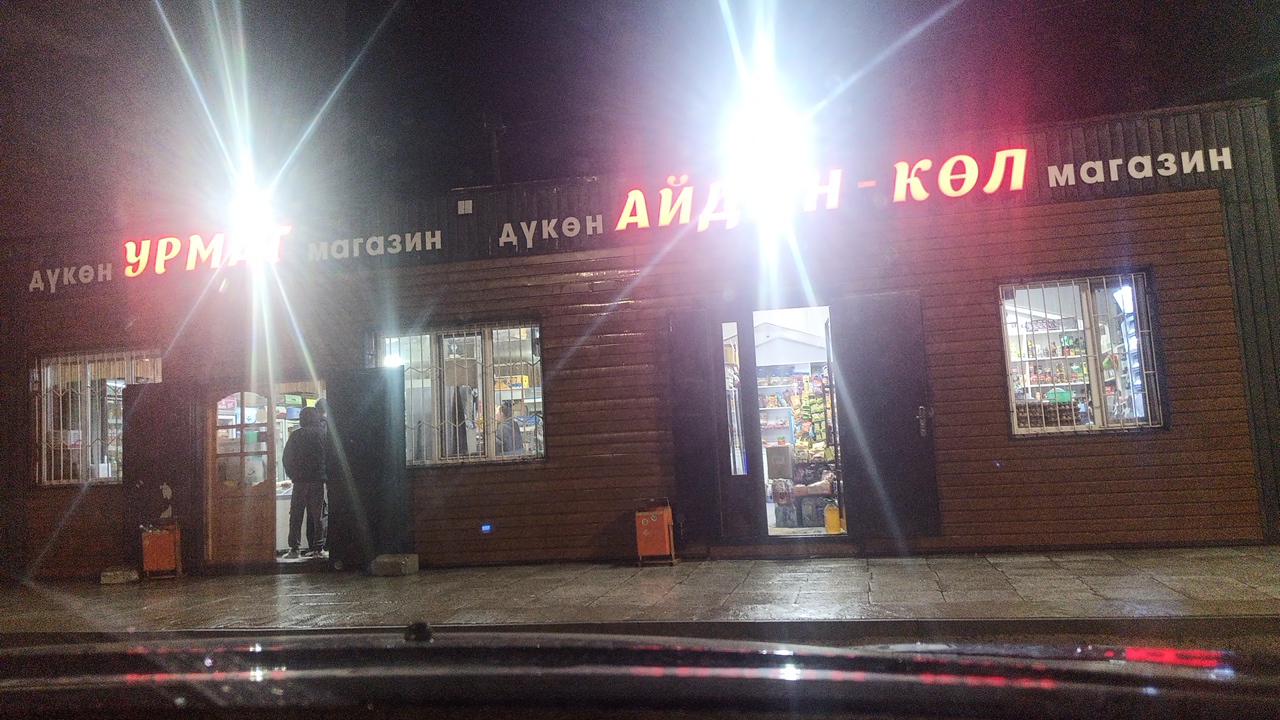 A nighttime shot shows the exterior of a store in Balykchy, Kyrgyzstan. The store has a wooden facade with several windows and two entrances. The main entrance is an open doorway leading into a brightly lit interior, revealing shelves stocked with goods. Another entrance is a closed dark door on the left. Two people are visible standing near the open doorway. The store has a prominent neon sign across the top. The text on the sign appears to read "ДҮКӨН УРМАТ" on the left and "ДҮКӨН АЙДАН - КӨЛ МАГАЗИН" on the right, indicating it is a store named "Urmat" and "Aidan-Kol Magazin". The scene is illuminated by bright lights from the store and from the neon sign, creating lens flares. The ground in front of the store appears wet, suggesting it may have rained recently. The overall atmosphere is that of a typical local convenience store operating in the evening.