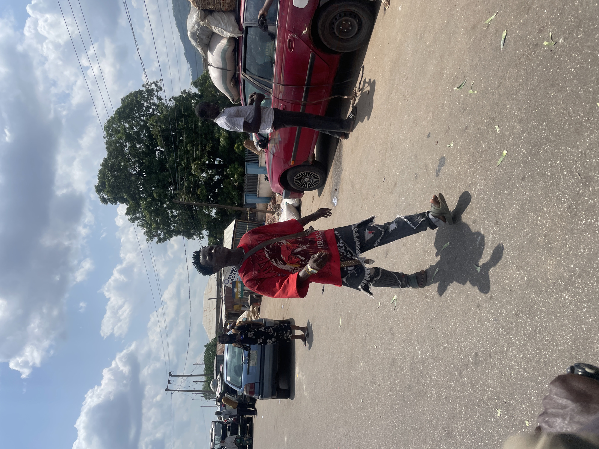 The image depicts a street scene in Lokoja, Nigeria. A young man in a red shirt and patterned jeans stands on the pavement. A red car laden with goods tied to its roof is parked nearby, where another man stands beside it. Other parked cars and motorcycles are visible in the background, with shops lining the street. The sky is cloudy, and there is a large tree nearby.