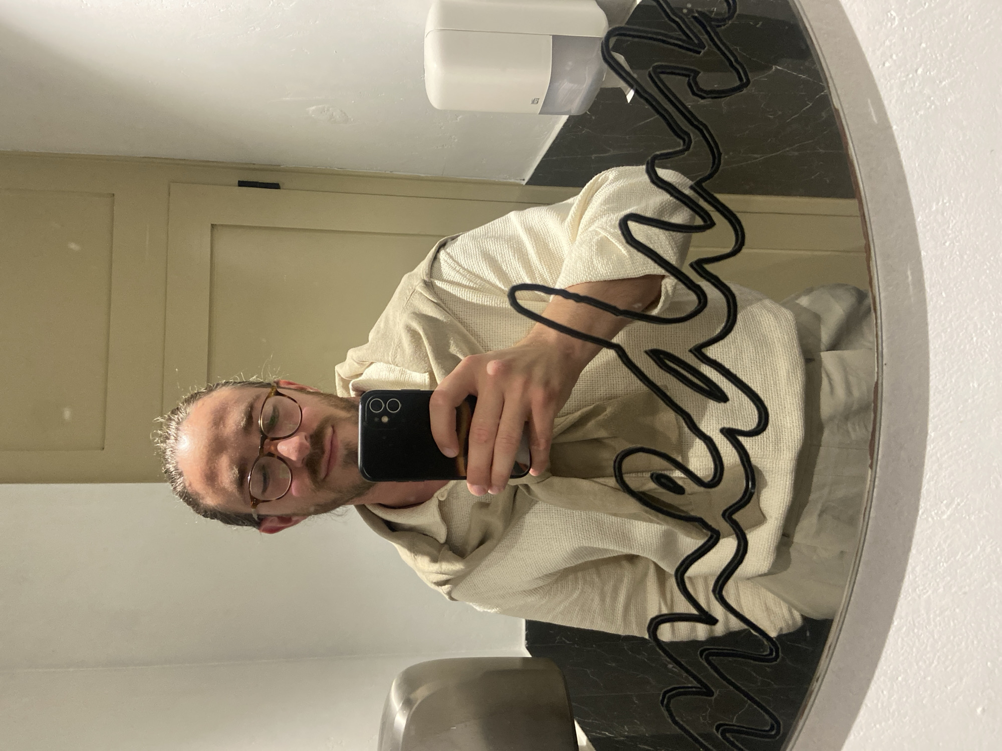 A man with glasses and a beige waffle-weave robe takes a selfie in a bathroom in Petrovac centar, Montenegro. The man is visible in the reflection of a round mirror with a decorative black squiggle design around its edge. A white soap dispenser is mounted above the mirror. The bathroom has white walls and beige doors, and black marble countertops.