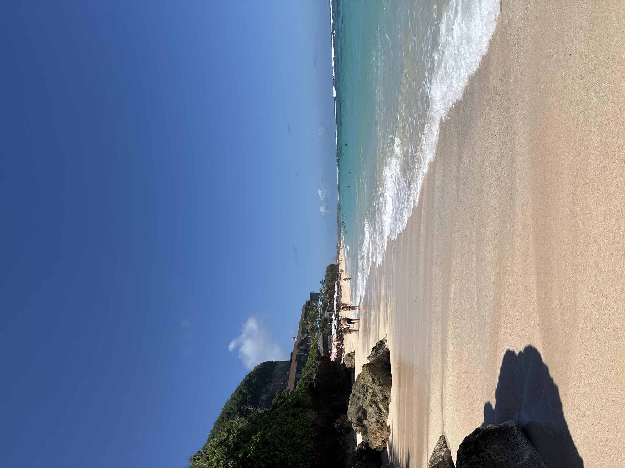 The image depicts a beach in Kuta Selatan, Indonesia, under a clear, blue sky. The scene is divided between the sky and the beach/ocean. The ocean has a turquoise hue and white waves are breaking onto the sandy shore. A line of people and beach umbrellas dot the shoreline. Beyond this, vegetation covers a rocky cliff. The foreground is filled with large rocks and dark shadows cast onto the sand.
