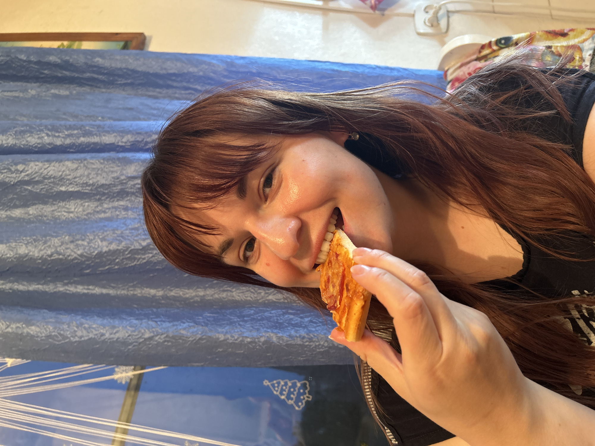 A young woman with brown hair is holding a slice of pizza to her mouth, apparently about to take a bite. The pizza has toppings of what appears to be cheese and a reddish sauce. She is smiling. Behind her is a blue backdrop, possibly a wall or curtain. In the background at the top, there is a white electrical outlet. The location cannot be determined from this picture.