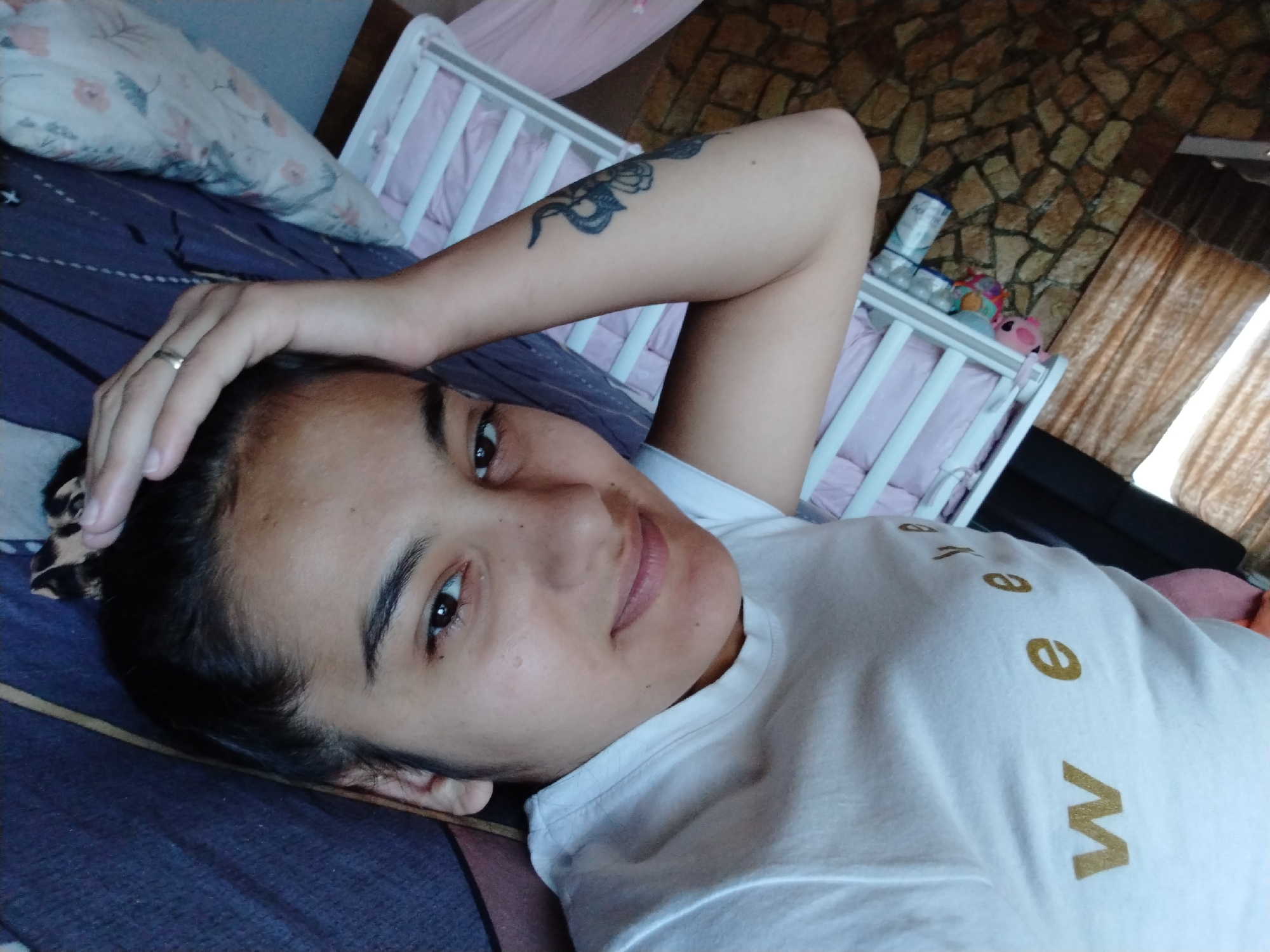 A woman with dark hair is lying on a bed with a blue and purple patterned duvet. She is wearing a white t-shirt with gold lettering. Her left arm is raised and her hand is resting on her head. She has a tattoo on her left arm. A gold ring is visible on her left hand. Behind her is a white crib with pink bedding. In the background, there is a stone wall and a window with gold curtains. A bottle and various toys are visible in the crib. The location in the city of Unknown, Unknown is unknown.